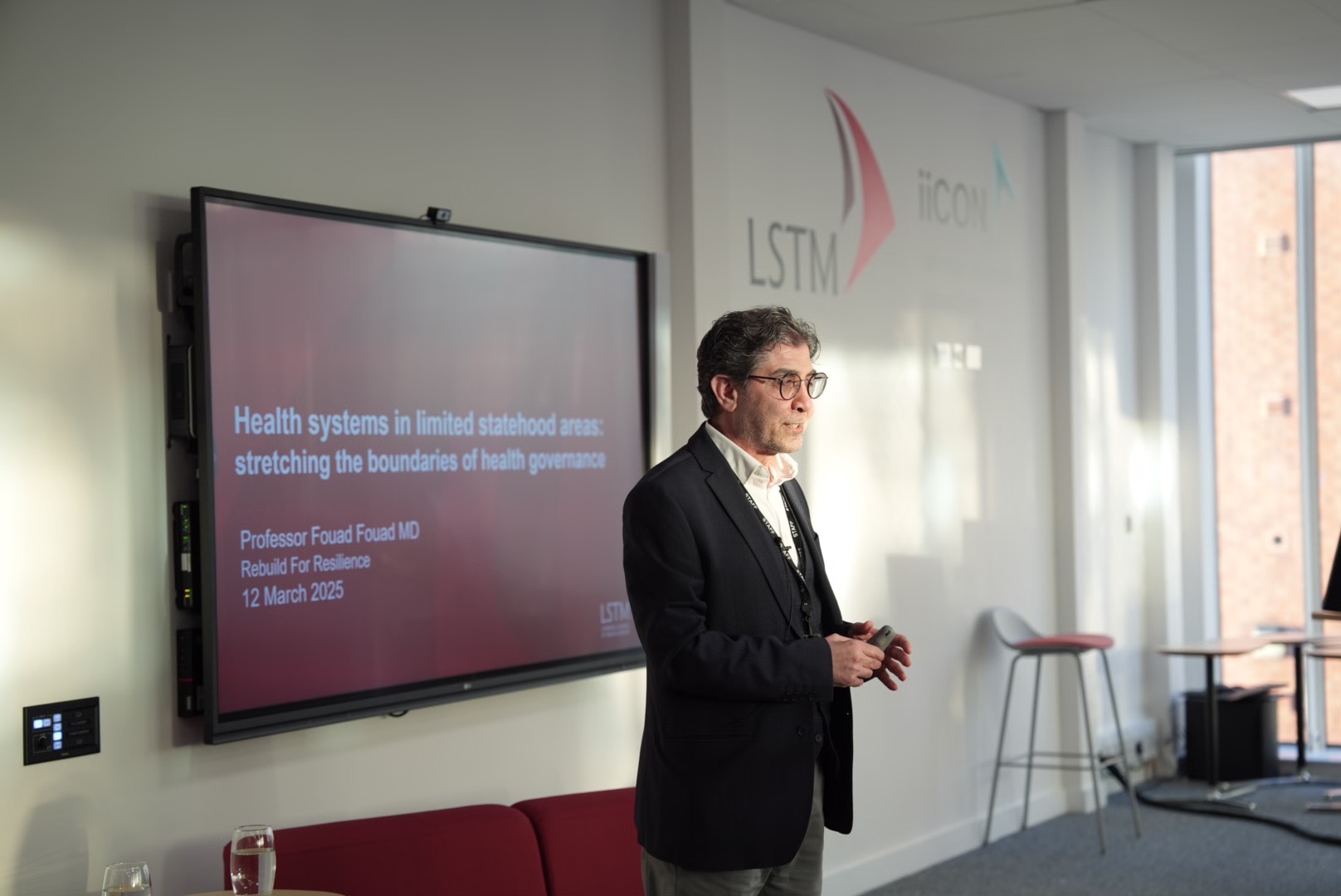 A lecturer speaks beside a large screen displaying a title slide reading “Health systems in limited statehood areas: stretching the boundaries of health governance”, with LSTM and iiCON logos on the wall behind.