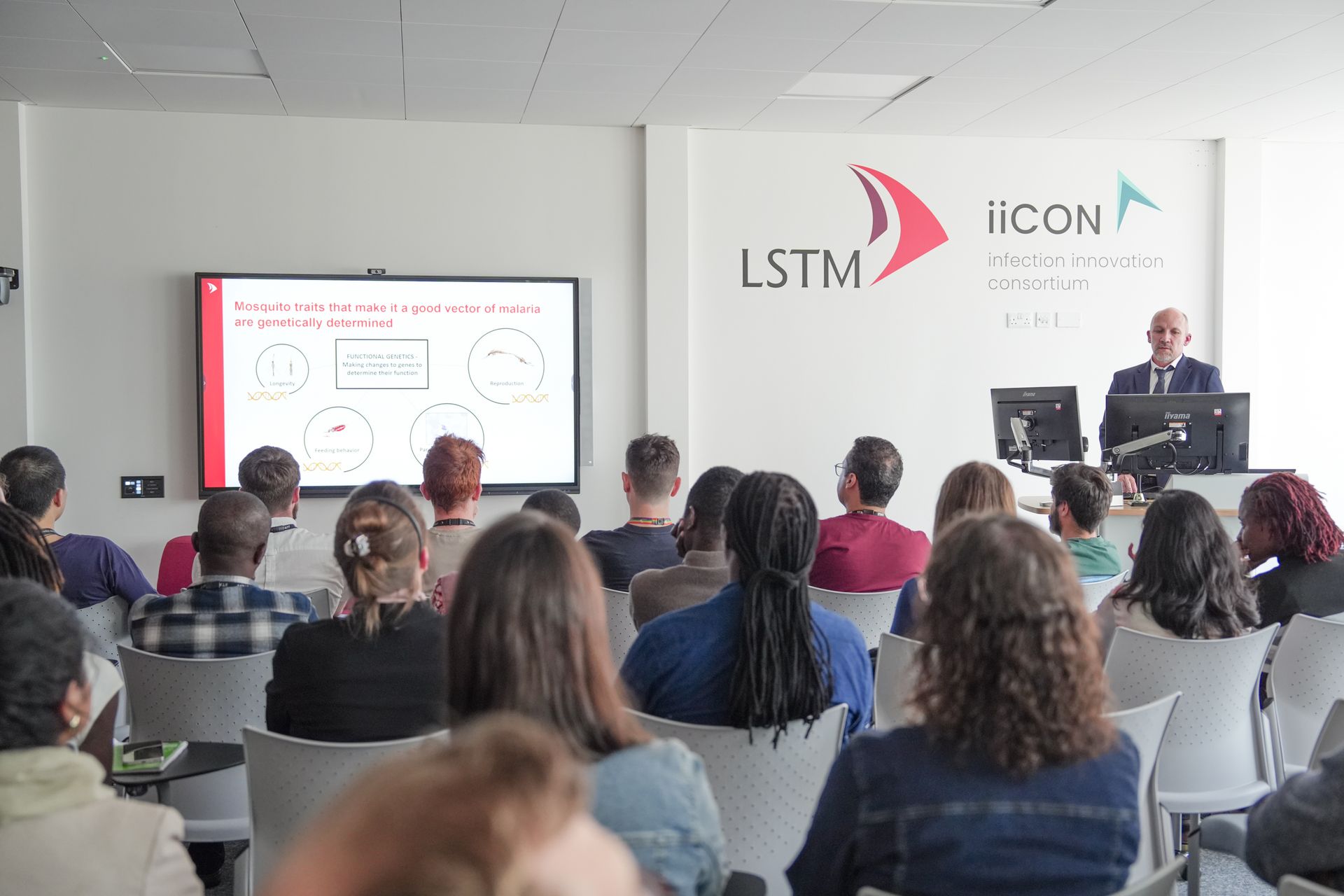 An audience listens to a lecture at LSTM as a speaker stands at the front beside a screen showing a slide on genetically determined mosquito traits linked to malaria transmission, with LSTM and iiCON logos on the wall behind.