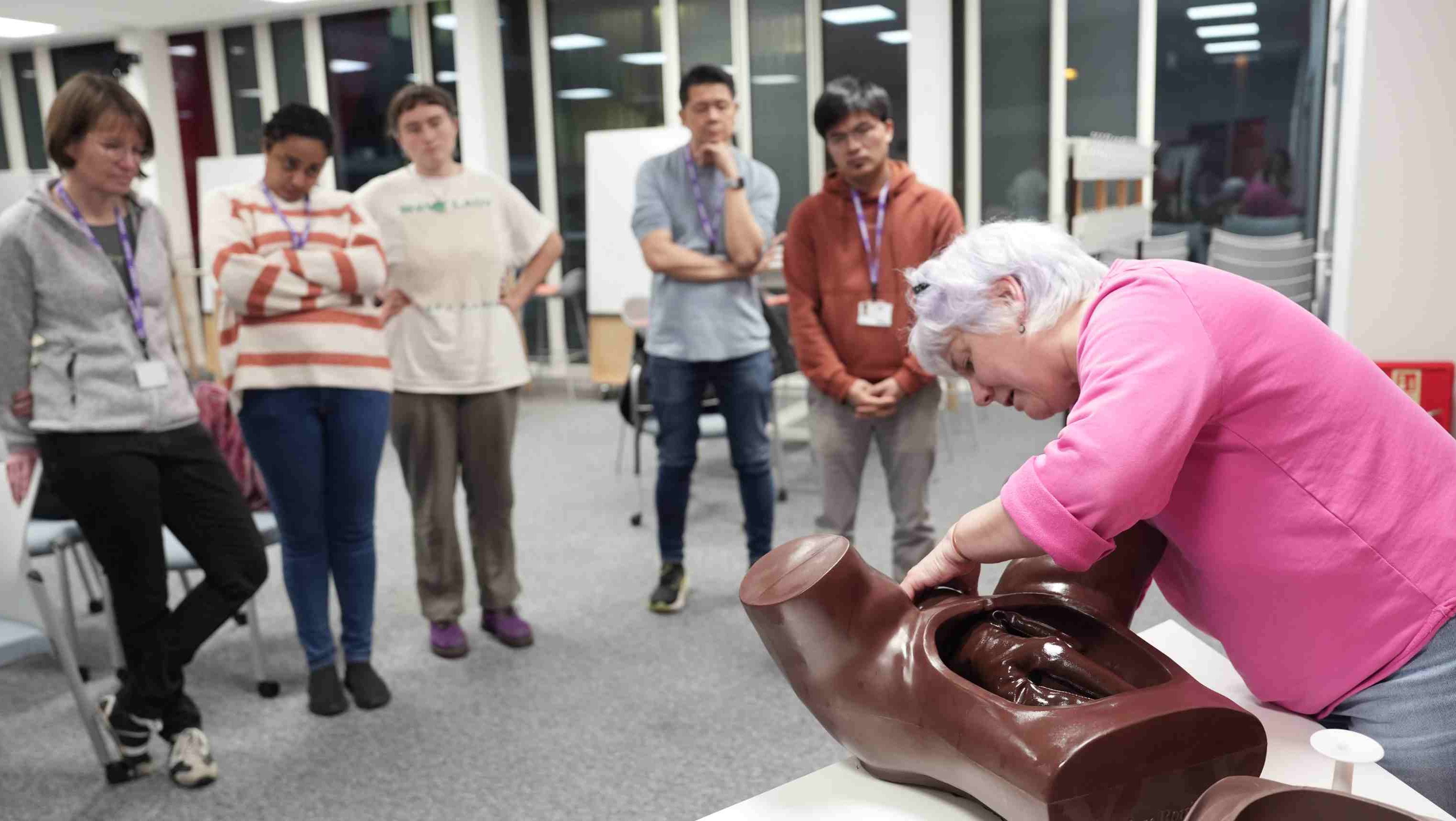 Trainer demonstrates a clinical skills technique on a medical training manikin while a group of learners watch in a classroom.
