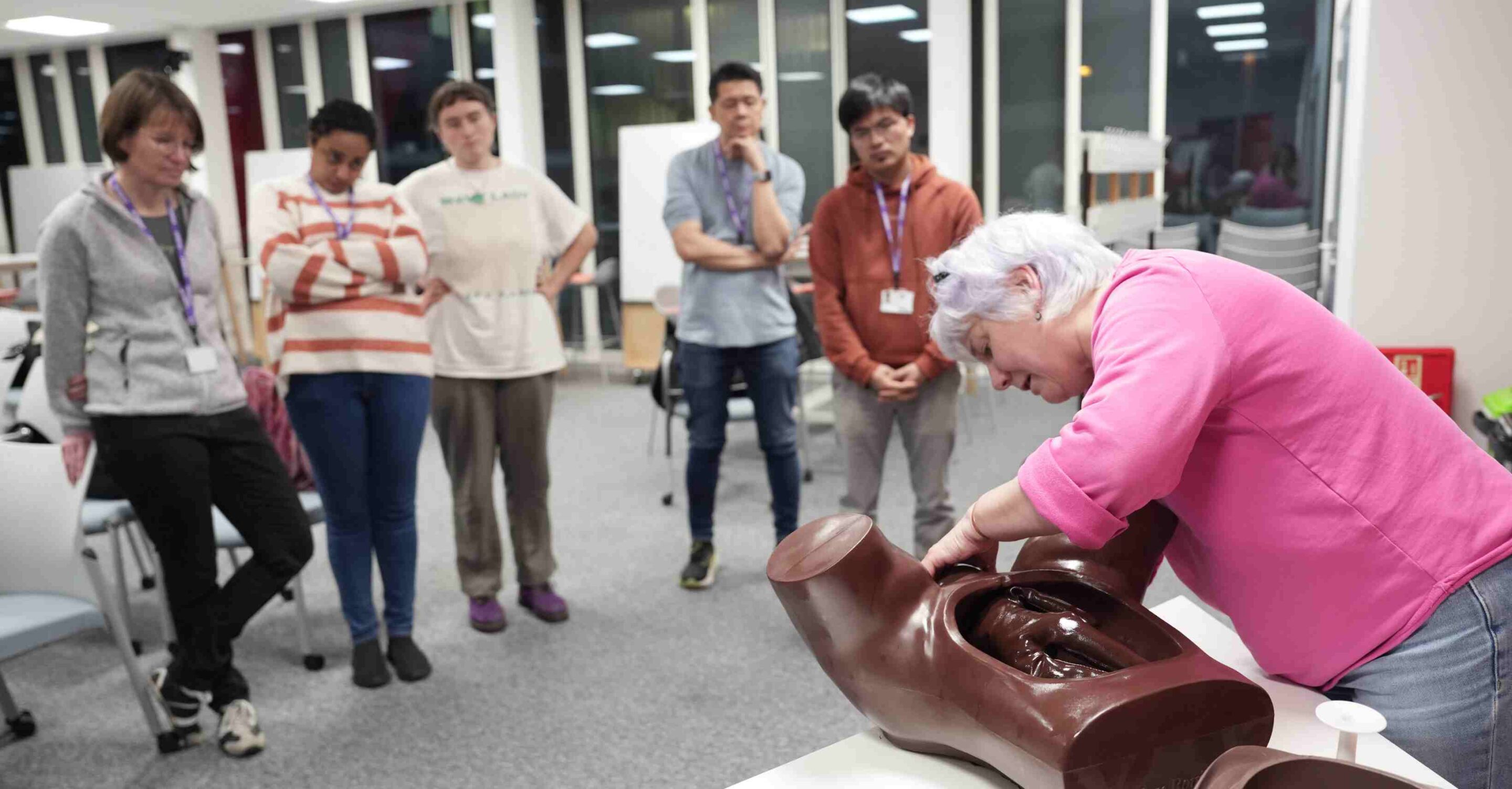 Trainer demonstrates a clinical skills technique on a medical training manikin while a group of learners watch in a classroom.
