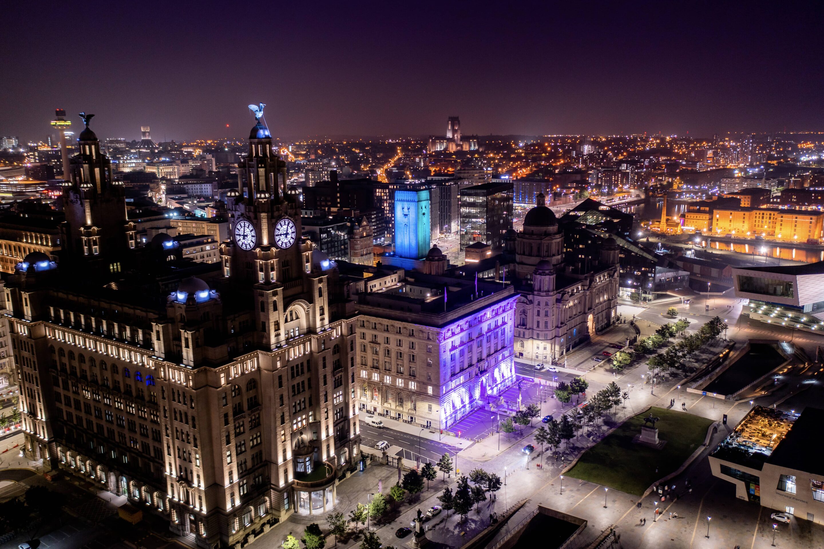 Aerial night view of Liverpool’s waterfront, with the illuminated Royal Liver Building and surrounding city lights stretching into the distance.
