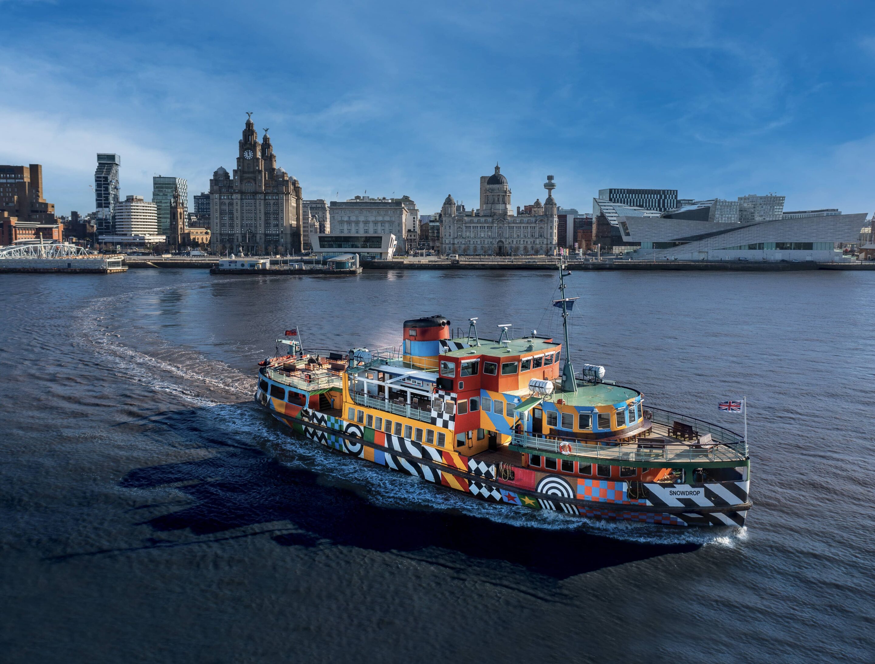 Colourful Mersey ferry crossing the River Mersey, with Liverpool’s waterfront skyline and the Royal Liver Building in the background.