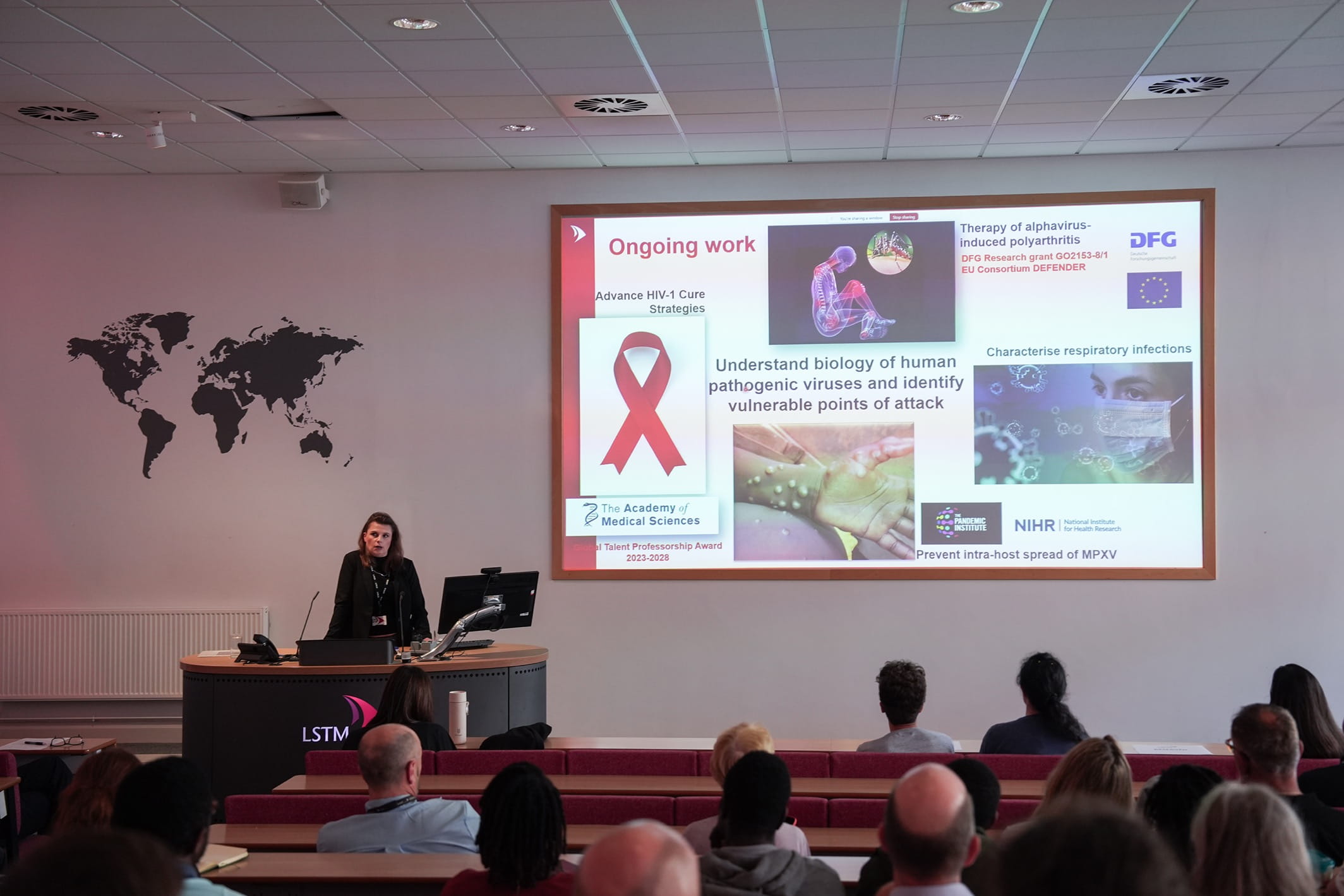A lecturer speaks at an LSTM podium to an audience in a lecture theatre, with a slide titled “Ongoing work” showing research on HIV cure strategies, pathogenic viruses, respiratory infections and MPXV.