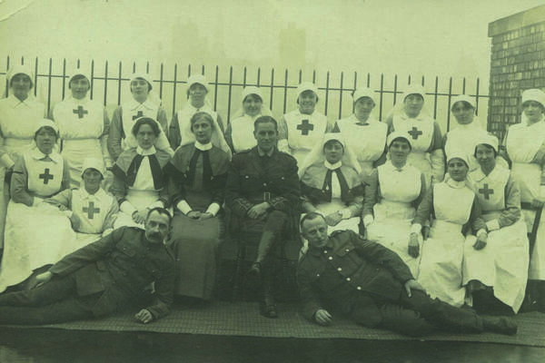 Vintage group photo of Red Cross nurses in uniform with two seated women and several soldiers, posed outdoors in front of a fence.