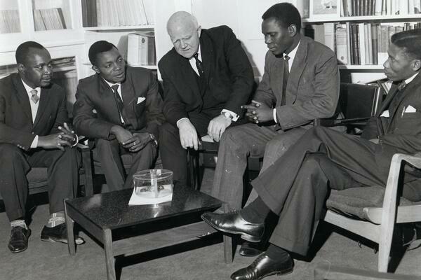 Black-and-white photo of five men in suits seated in a meeting room, talking around a low table with papers, with bookshelves behind them.