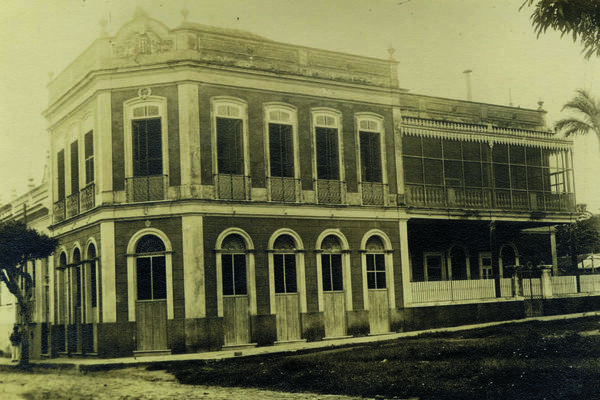 Sepia-toned historic photo of a two-storey colonial-style building with arched windows and a wraparound balcony.