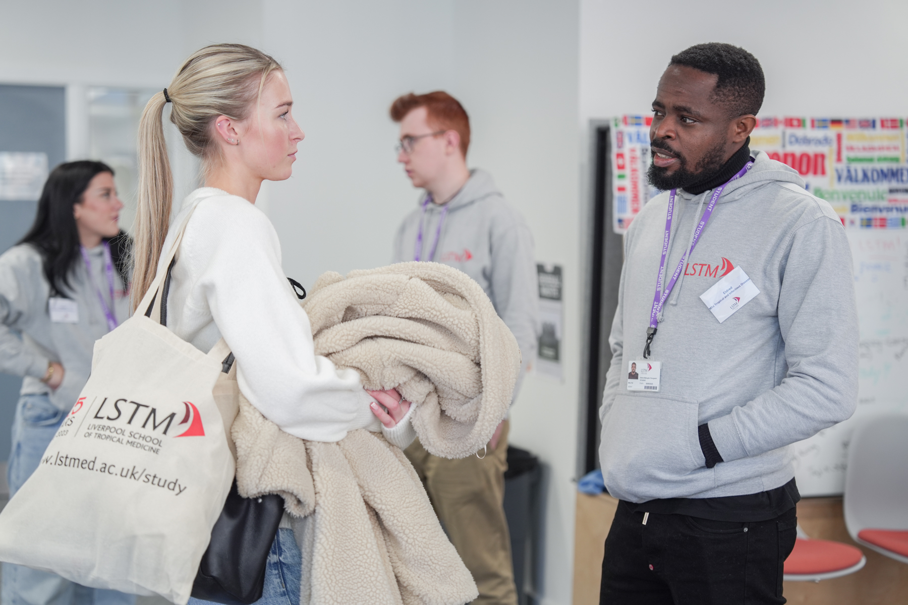 Two people talking at an LSTM event or open day, with both wearing lanyards and LSTM branding visible on a hoodie and tote bag.