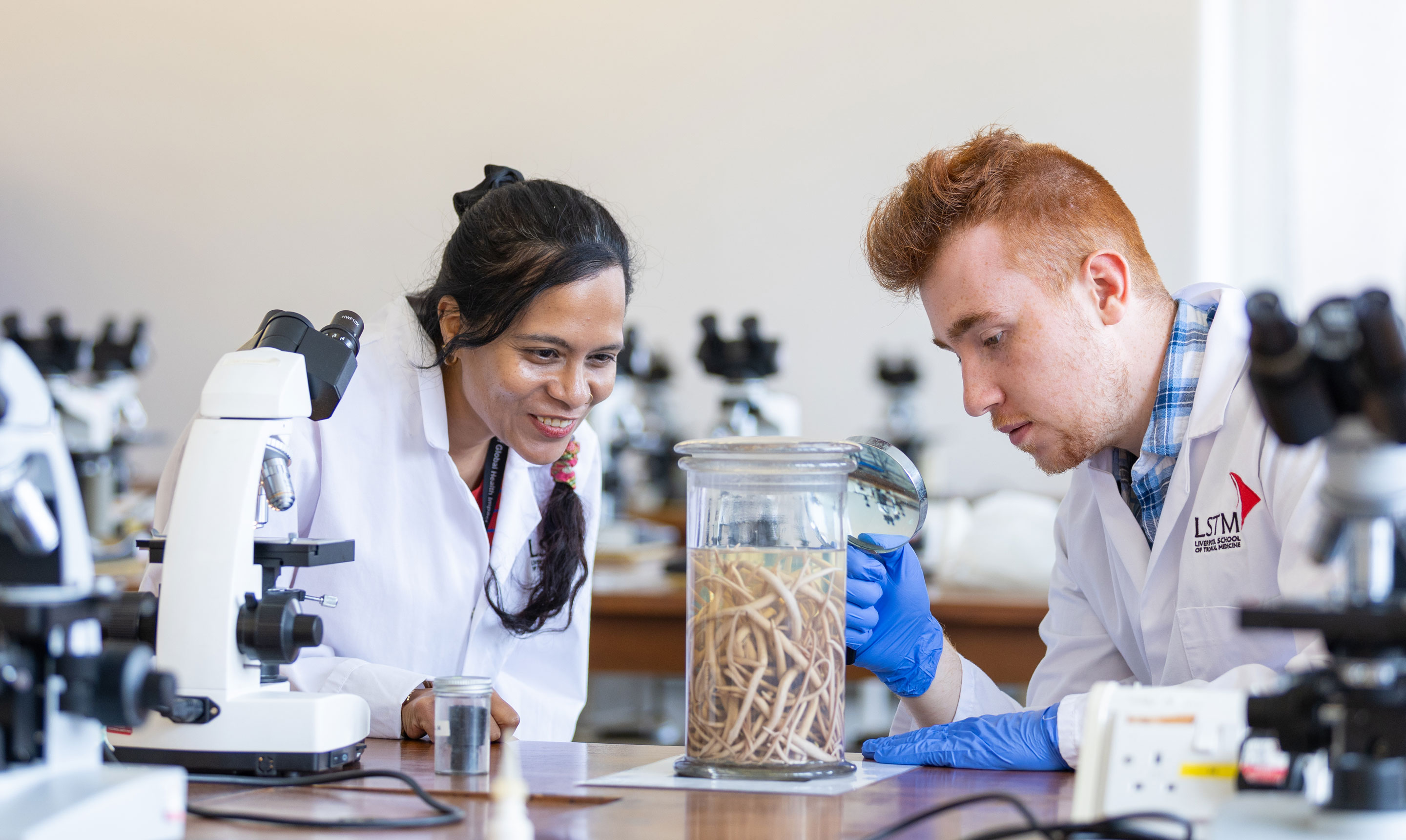 Two postgraduate students in white LSTM lab coats examine a specimen jar containing preserved worms, using microscopes and lab equipment in a teaching laboratory.