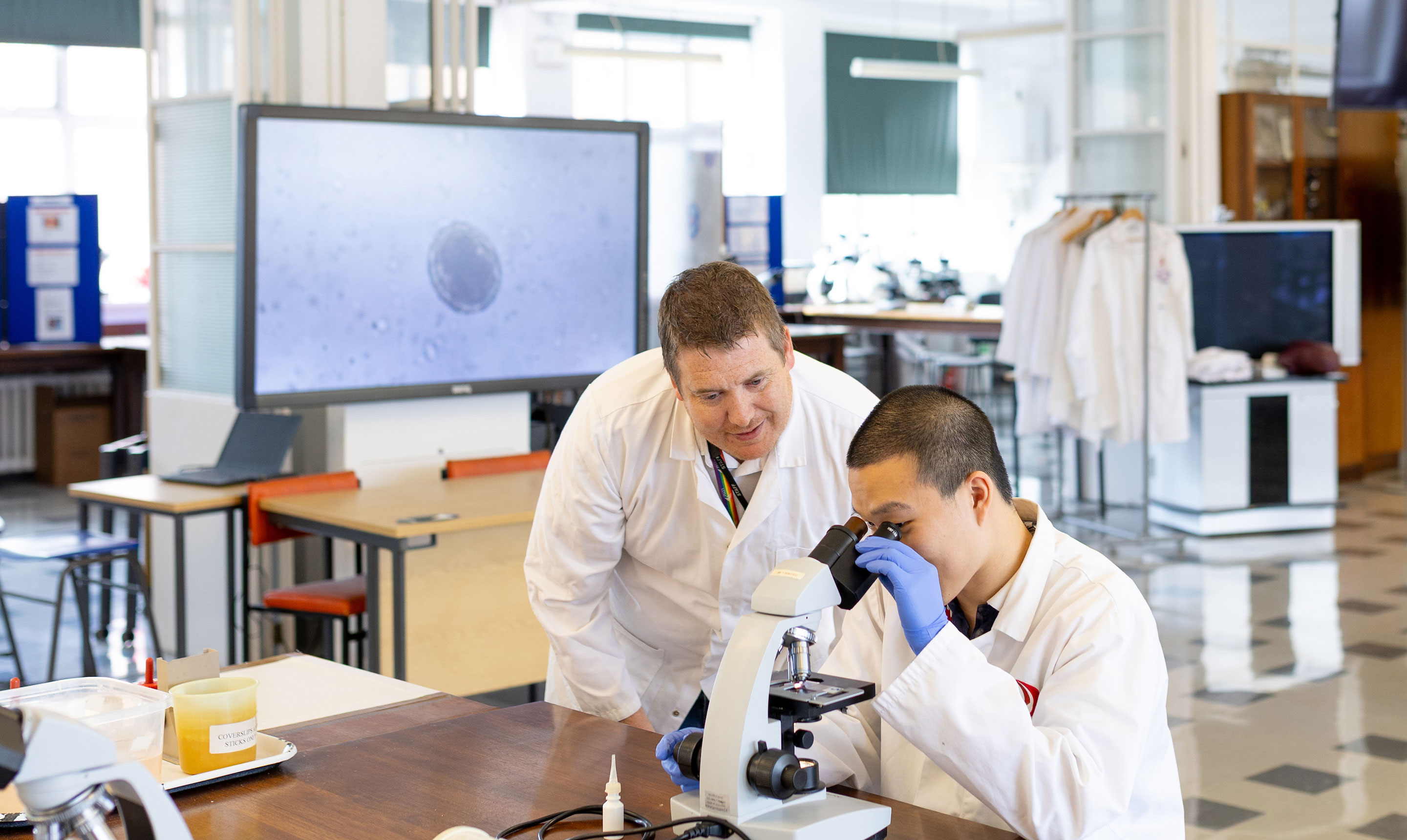 A student in a white lab coat and blue gloves looks through a microscope at a laboratory bench, while a teacher in a white lab coat leans over to observe and support. On the bench are lab items including a plastic container, labelled tape, and a dropper. In the background, a large digital screen displays a magnified microscopic image, and several lab coats hang on a rack. The bright, modern laboratory is equipped with desks, stools, and additional microscopes.