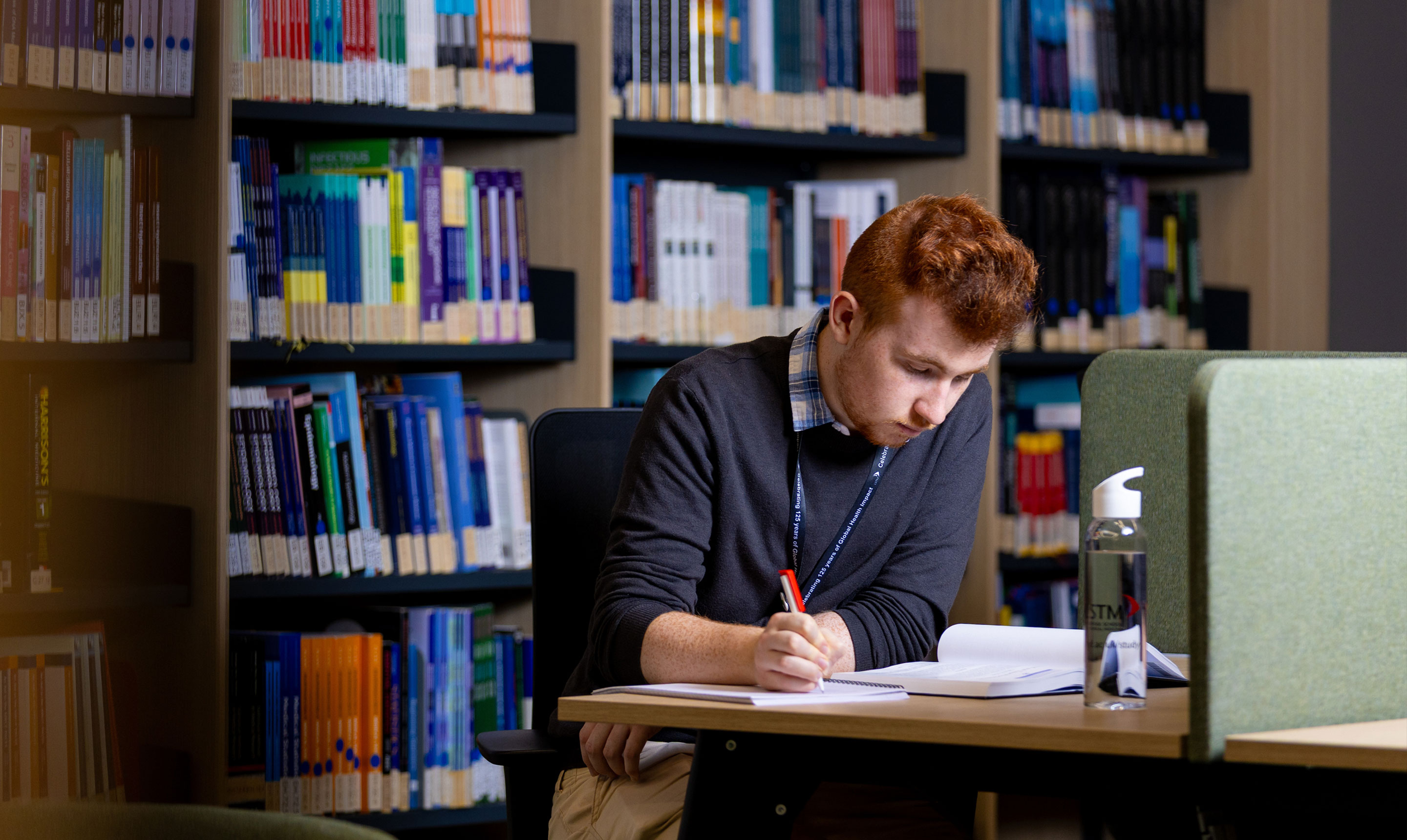 A student with red hair sits at a desk in a library, focused on writing notes in a notebook with a red pen. He wears a dark jumper over a checkered shirt and a lanyard around his neck. Shelves filled with colourful academic books line the background. On the desk in front of him is an open notebook, and a clear water bottle with the Liverpool School of Tropical Medicine (LSTM) logo. Green fabric study dividers provide a quiet, focused workspace.