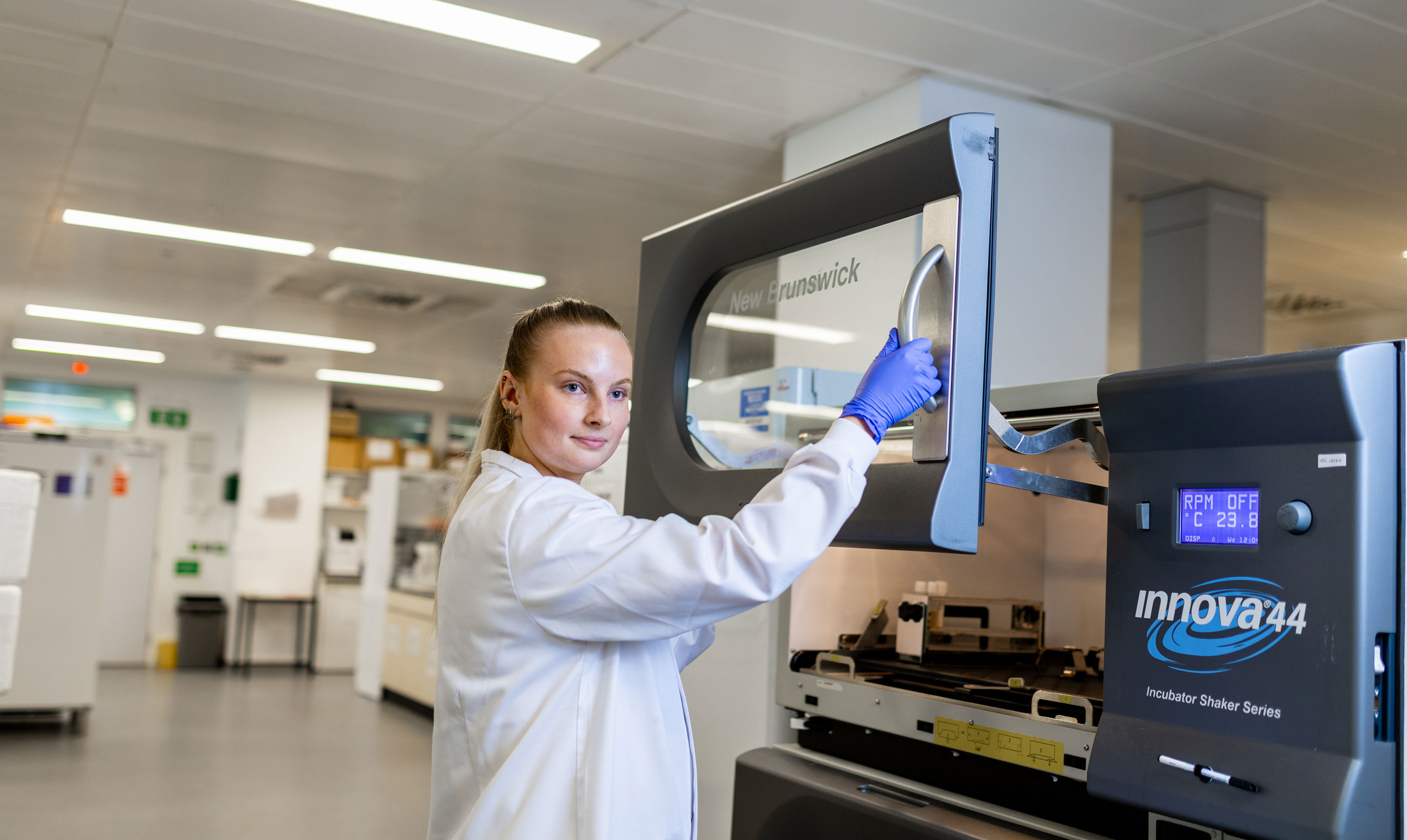 A female researcher wearing a white lab coat and blue protective gloves opens the door of a large laboratory incubator shaker, labelled New Brunswick Innova 44. She looks towards the camera while handling the equipment, which displays a digital screen showing temperature and RPM settings. The surrounding lab space is modern and well-lit, with benches, storage units, and laboratory equipment visible in the background.