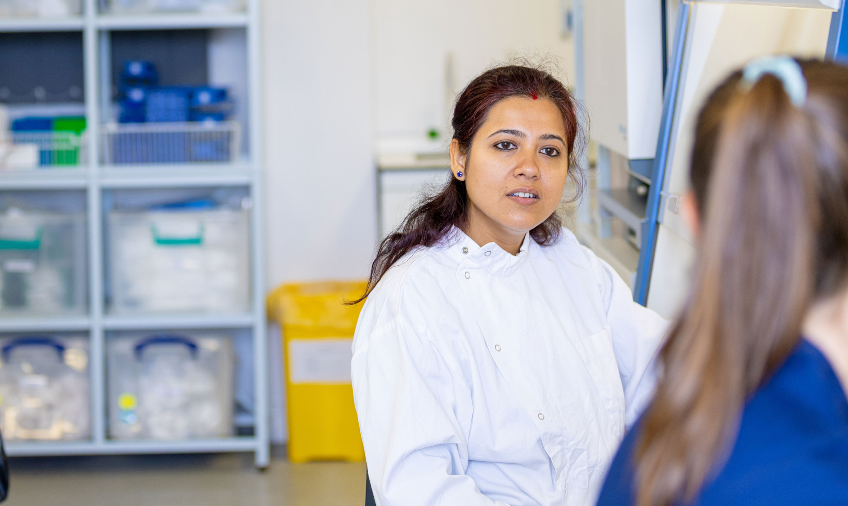 A female researcher wearing a white laboratory coat is engaged in conversation with a colleague inside a lab. She has dark hair tied back and is facing slightly towards the camera while speaking. In the foreground, the blurred figure of another person with long hair tied in a ponytail is visible, listening. Behind them, shelves stacked with plastic storage containers and a yellow clinical waste bin suggest an active laboratory environment.