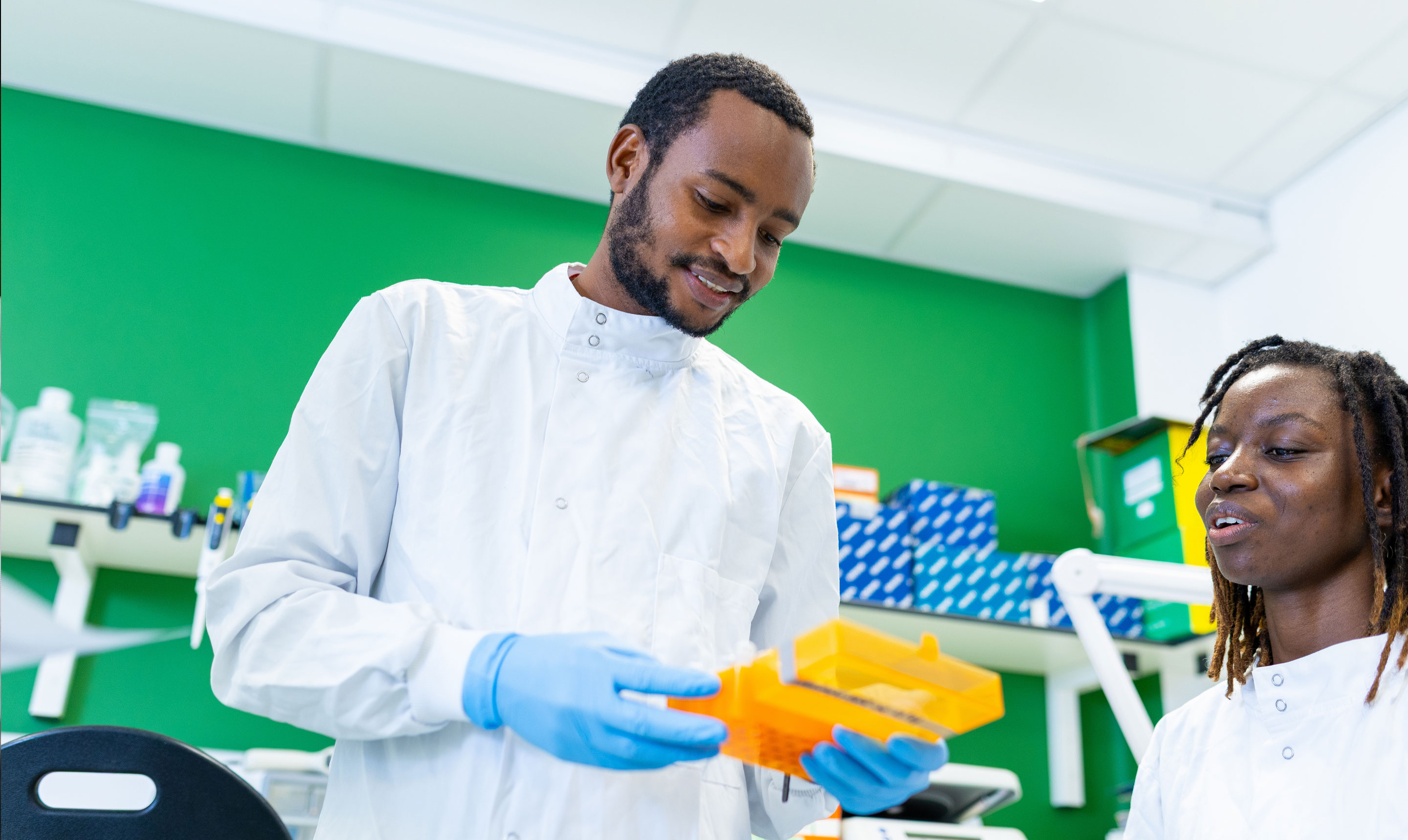 Two researchers in white lab coats and blue gloves work together in a brightly lit laboratory with green walls. One researcher holds a yellow plastic rack containing test tubes and smiles as he looks at it, while the other, seated nearby, is engaged in conversation. Behind them, laboratory shelves hold boxes, pipettes, and other equipment, creating a collaborative and professional research setting.