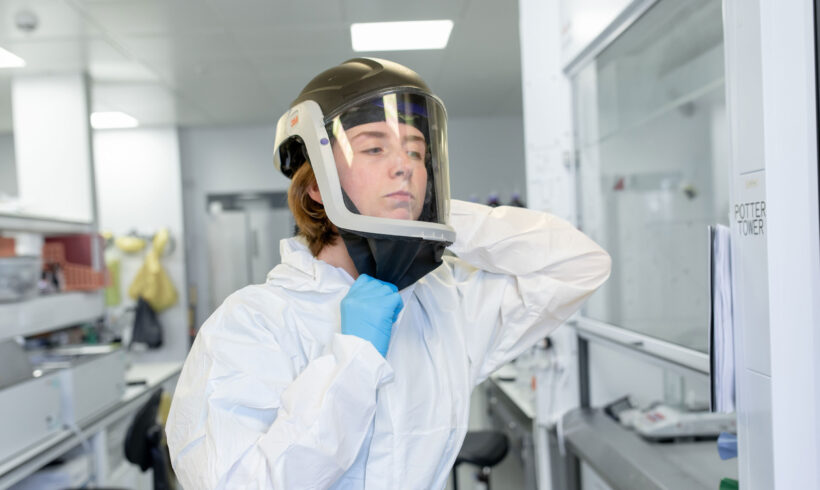 A researcher in a laboratory puts on full protective equipment, adjusting a helmet with a clear face shield while wearing a white disposable coverall suit and blue gloves. The individual is securing the lower edge of the helmet for a proper fit before entering or working in a controlled environment. The background shows a modern lab with benches, equipment, and safety signage, emphasising preparation for high-containment or hazardous material research.