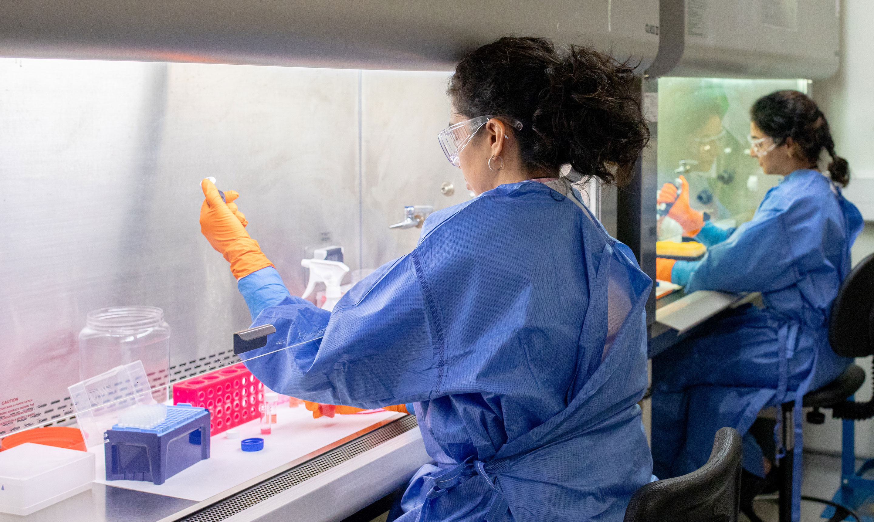 Two researchers wearing blue protective gowns, orange gloves, and safety goggles work at biosafety cabinets in a laboratory. The researcher in the foreground is holding a sample tube with one hand while using a pipette with the other, surrounded by racks of test tubes and laboratory containers. In the background, the second researcher mirrors the process at a separate workstation, highlighting parallel sample handling and sterile technique in a controlled environment.