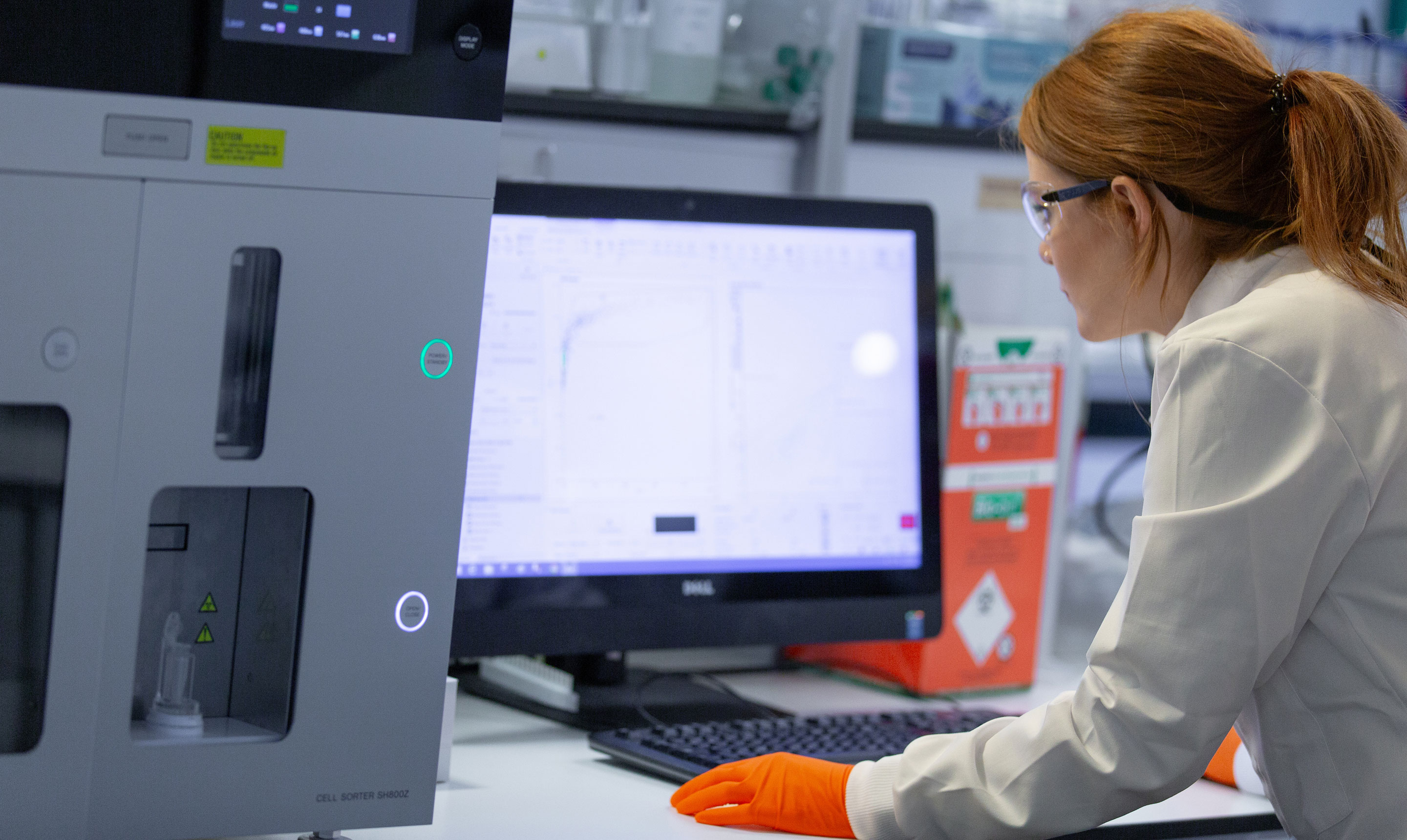 A female researcher in a white lab coat and orange protective gloves works at a computer connected to advanced laboratory equipment. She leans forward, closely examining scientific data displayed on the computer monitor, which shows charts and graphs from a cell analysis or sample processing system. The large laboratory machine beside her is labelled Cell Sorter SH800Z. Safety goggles, lab containers, and equipment are visible around the workstation, highlighting a high-tech biomedical research environment.