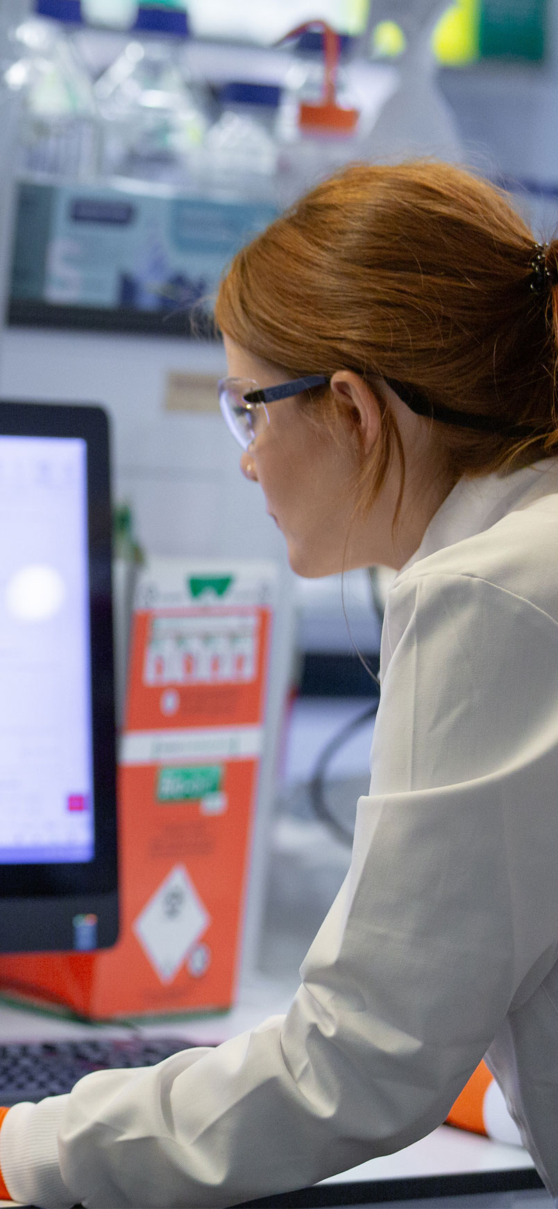 A female researcher in a white lab coat and orange protective gloves works at a computer connected to advanced laboratory equipment. She leans forward, closely examining scientific data displayed on the computer monitor, which shows charts and graphs from a cell analysis or sample processing system. The large laboratory machine beside her is labelled Cell Sorter SH800Z. Safety goggles, lab containers, and equipment are visible around the workstation, highlighting a high-tech biomedical research environment.