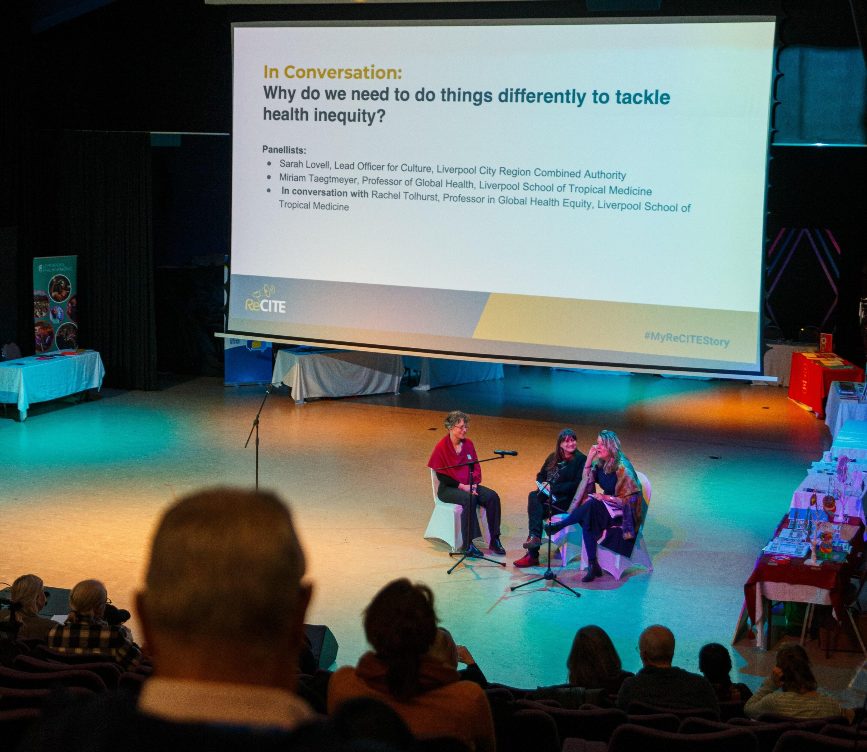 A photo taken from within an audience of people watching three academics on stage. They are talking and sat in front of a large presentation.