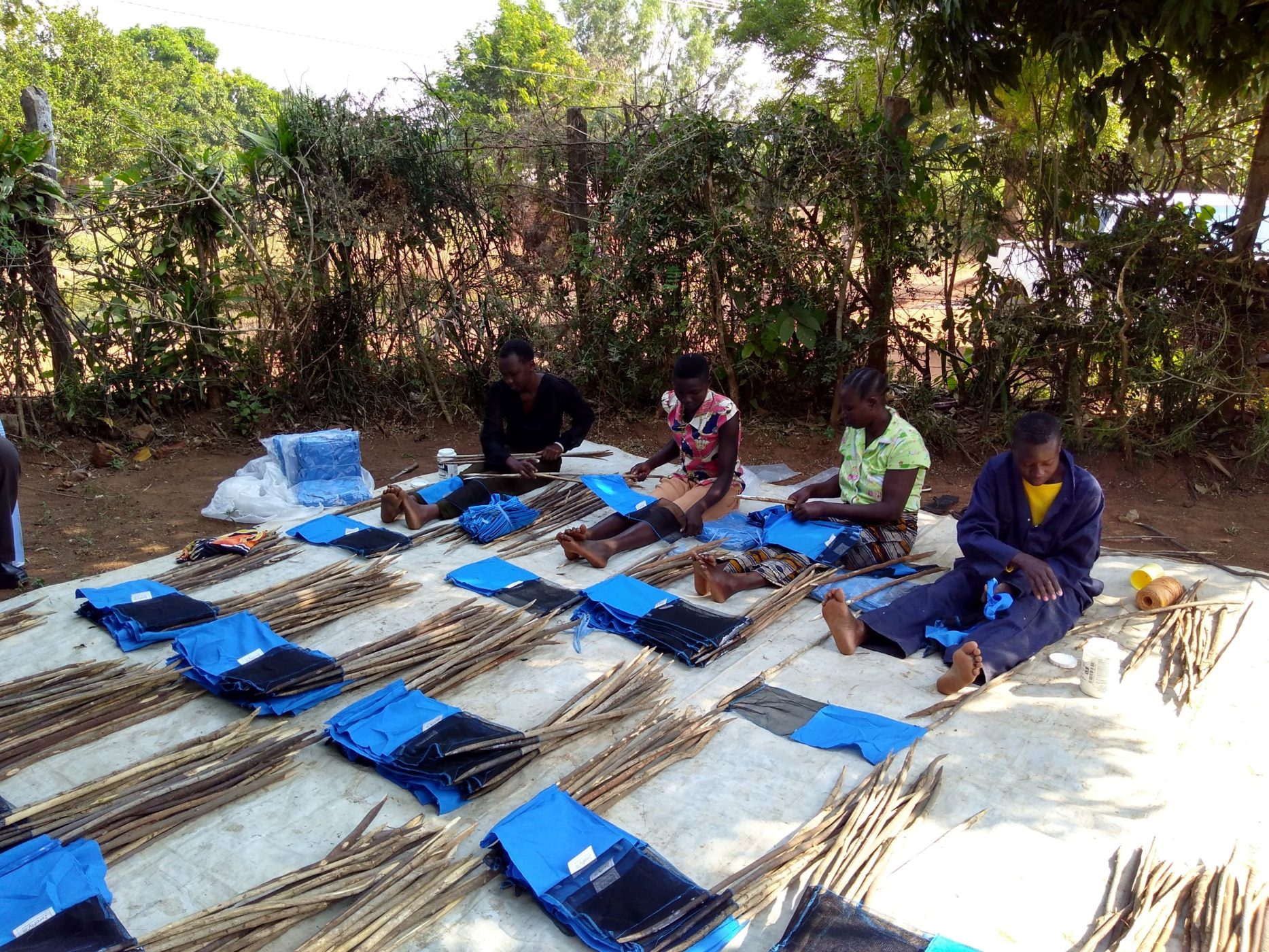 A photo of four people sat on a white mat putting together blue tiny targets, a kind of insect trap.