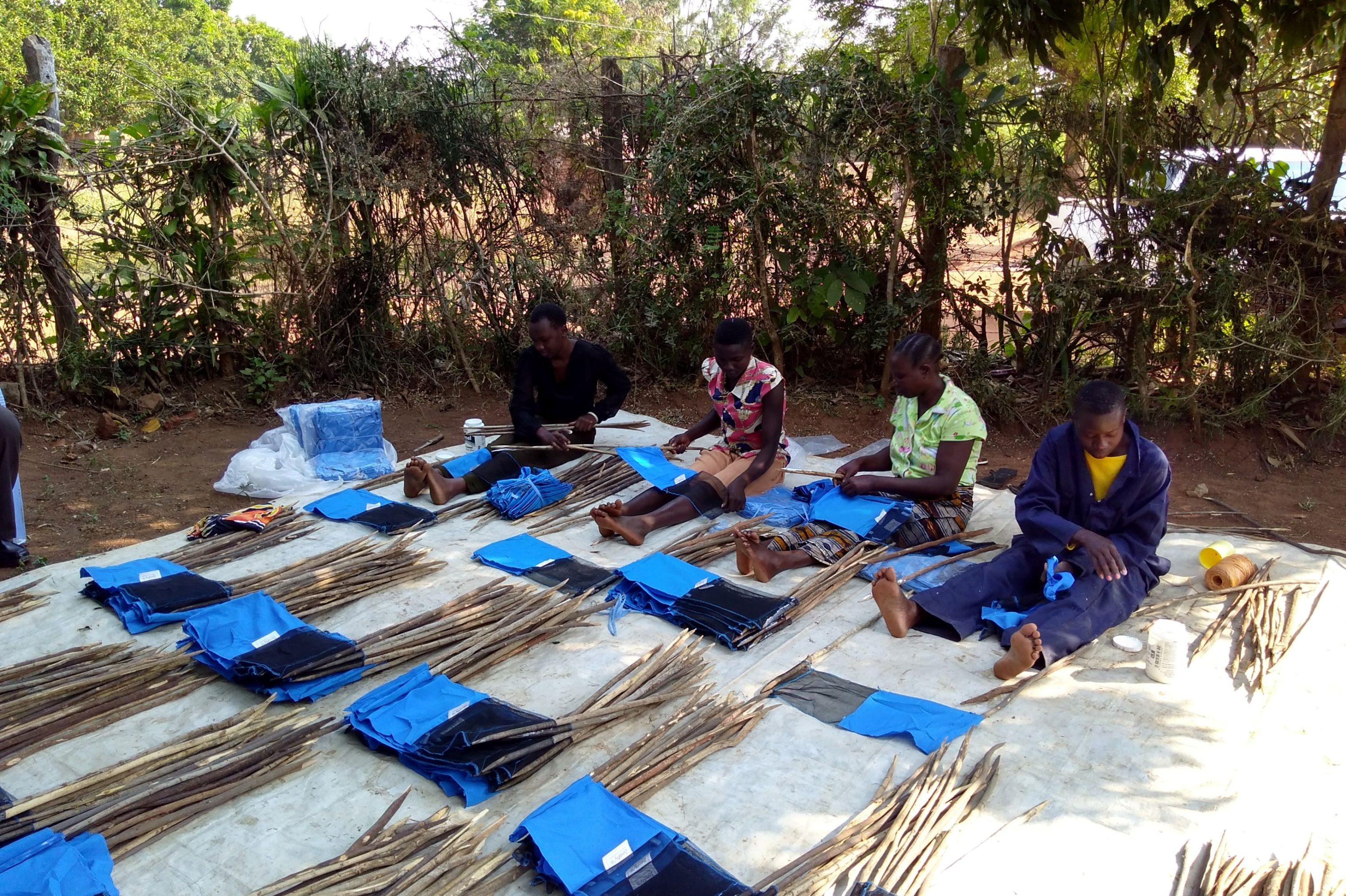 A photo of four people sat on a white mat putting together blue tiny targets, a kind of insect trap.