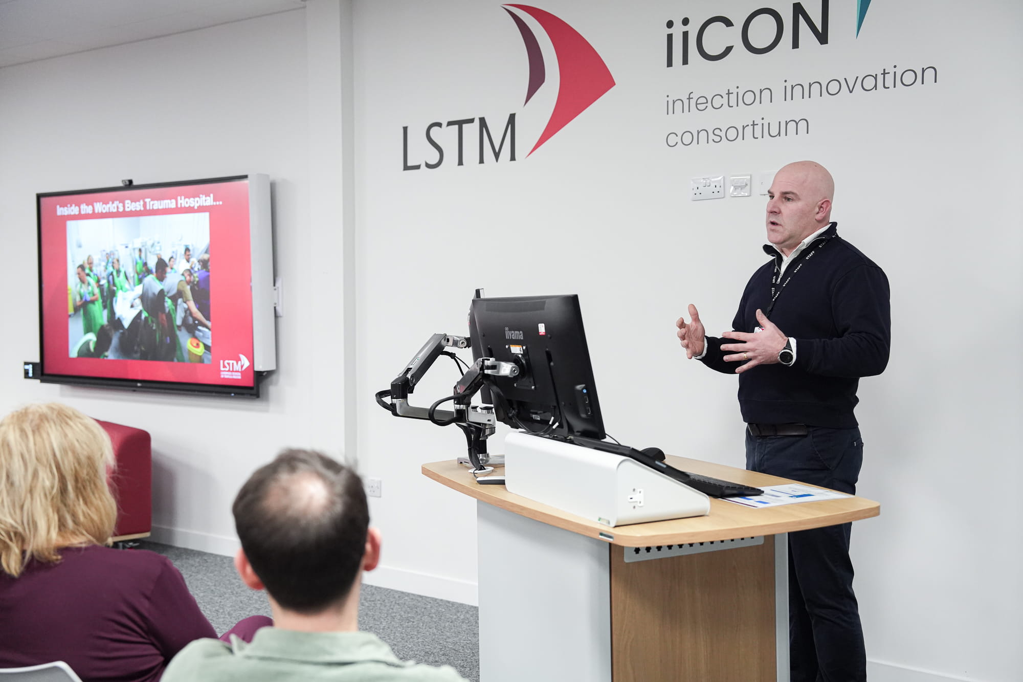 A speaker delivers a lecture at LSTM, standing beside a podium as a slide titled “Inside the World’s Best Trauma Hospital…” is shown on a screen, with the LSTM and iiCON (Infection Innovation Consortium) logos on the wall and audience members in the foreground.