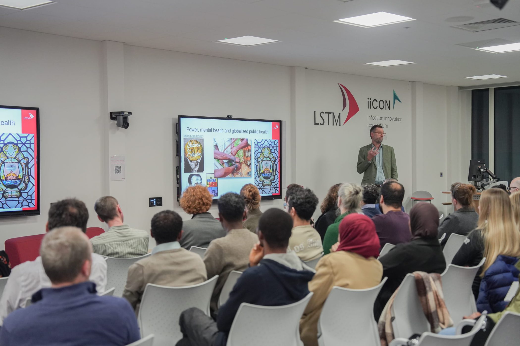 Audience members sit facing a speaker giving a lecture at LSTM, with a slide titled “Power, mental health and globalised public health” displayed on a screen and LSTM and iiCON logos on the wall behind.