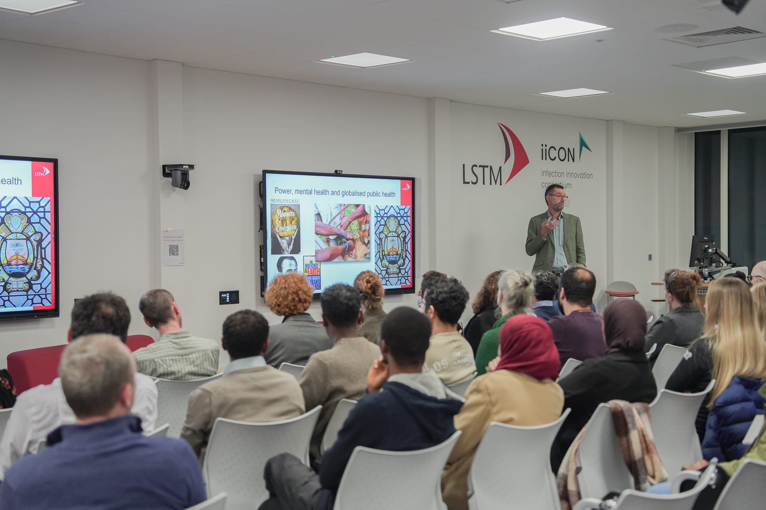 Audience members sit facing a speaker giving a lecture at LSTM, with a slide titled “Power, mental health and globalised public health” displayed on a screen and LSTM and iiCON logos on the wall behind.