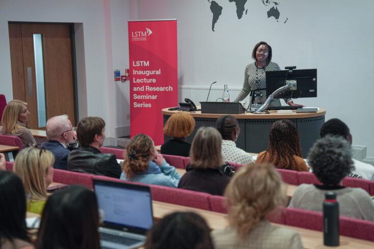 A speaker presents at the LSTM Inaugural Lecture and Research Seminar Series, standing at a lectern in front of an audience seated in a lecture theatre, with a red LSTM event banner beside the stage.