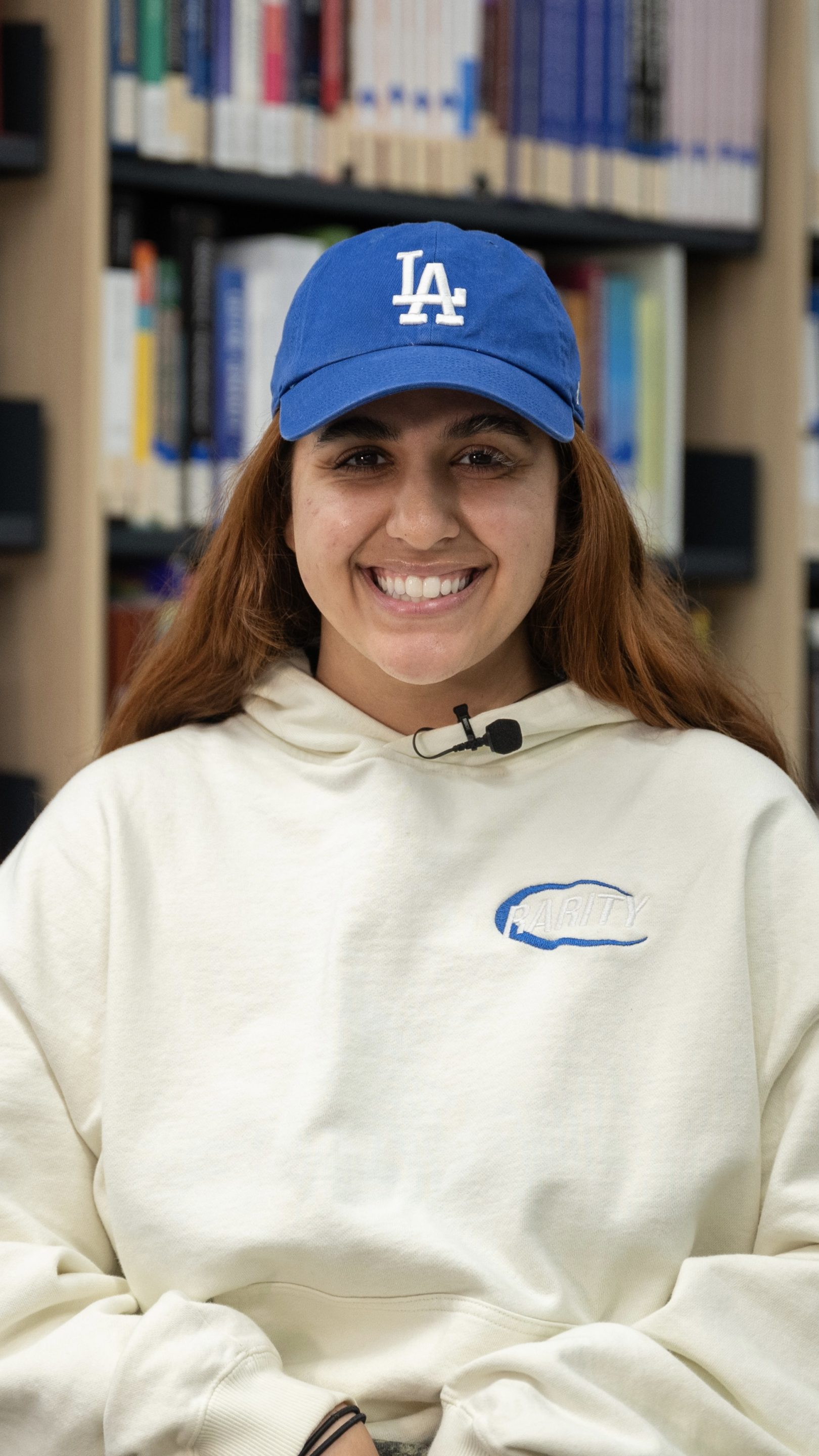 Person smiling while seated in a library, wearing a cream hoodie and a blue cap, with rows of books in the background.
