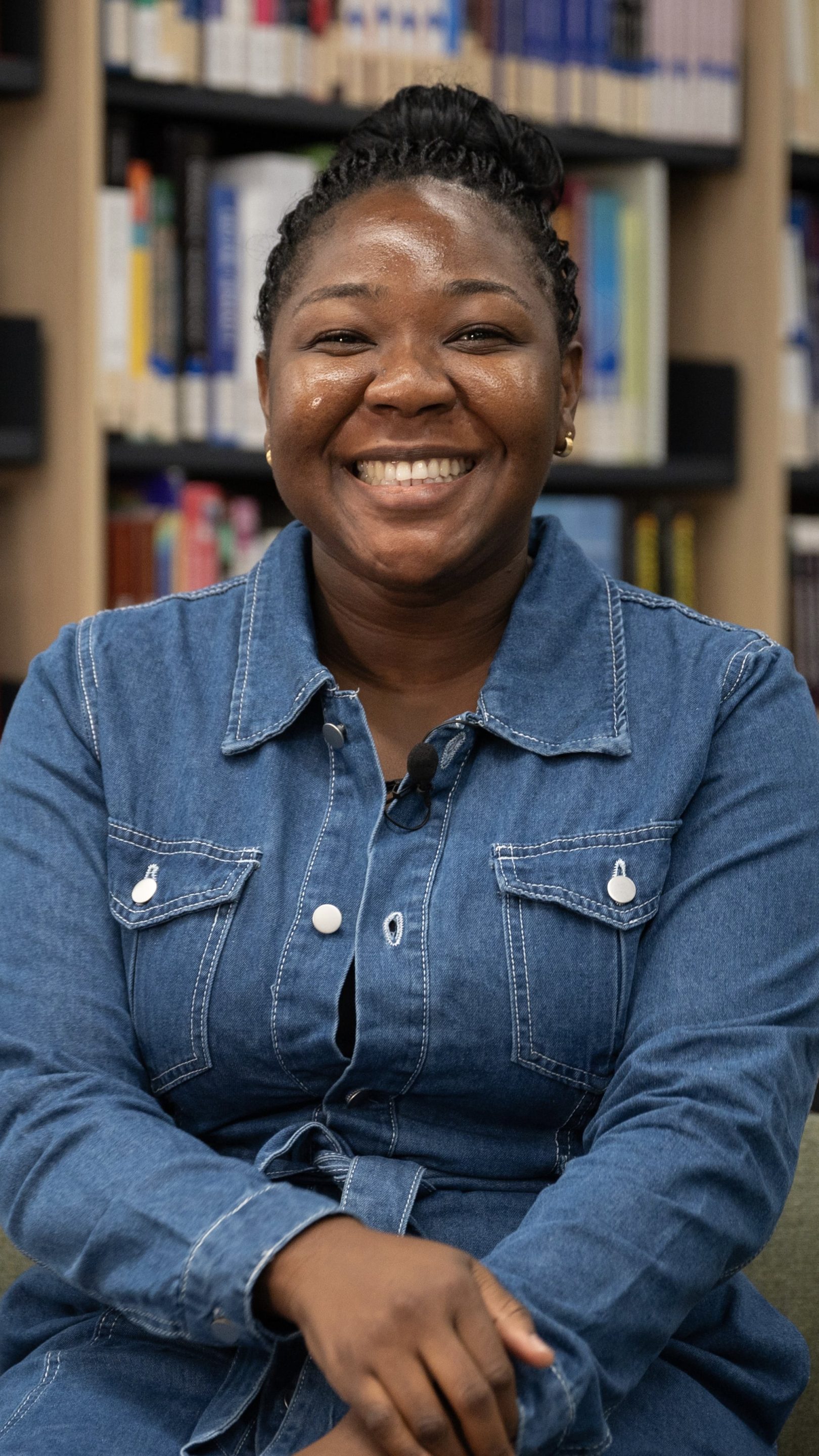 Person smiling and seated in a library, wearing a denim dress, with bookshelves visible behind them.