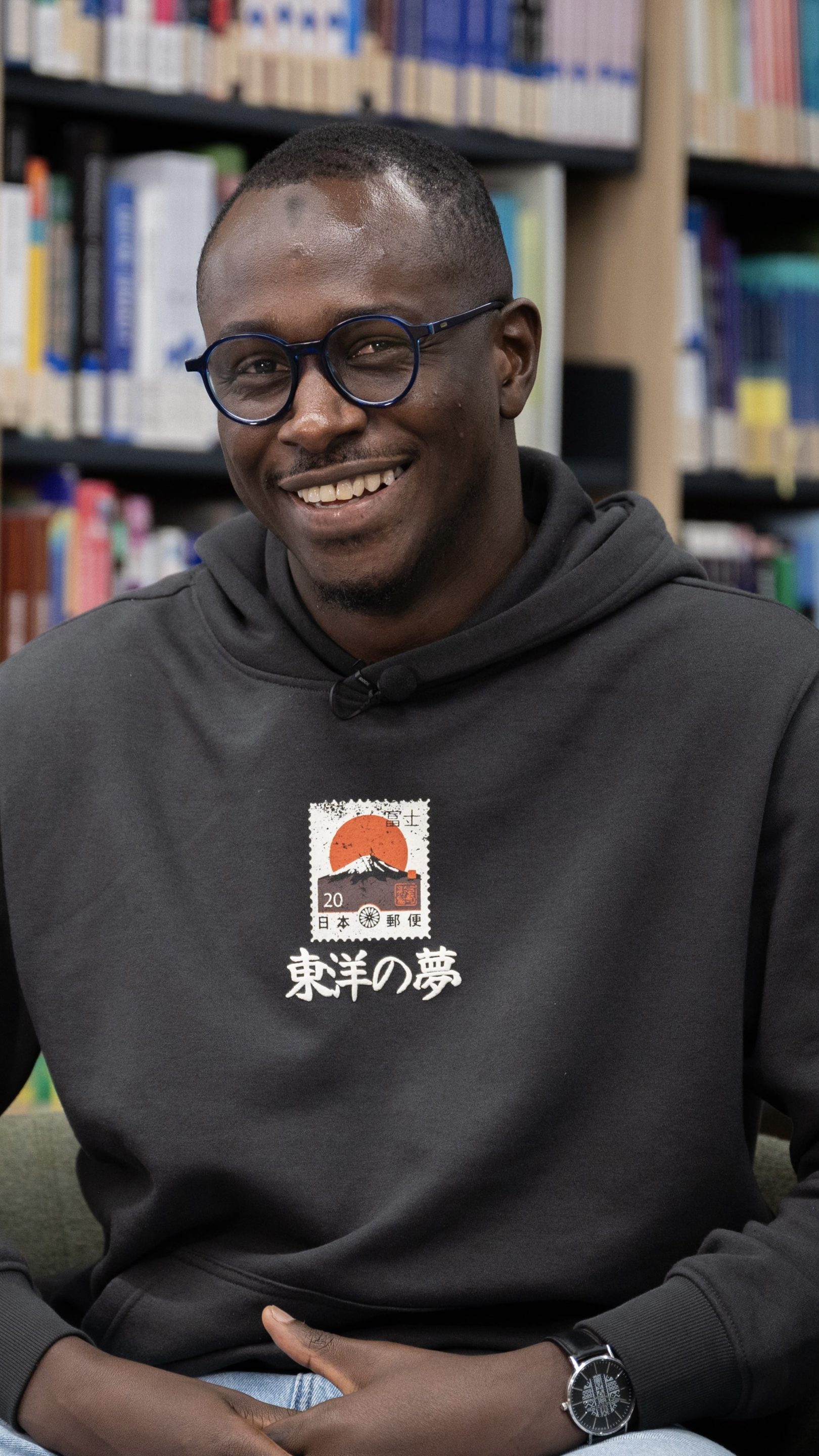 Person smiling while seated in a library, wearing a dark hoodie with a printed design and glasses, surrounded by bookshelves.