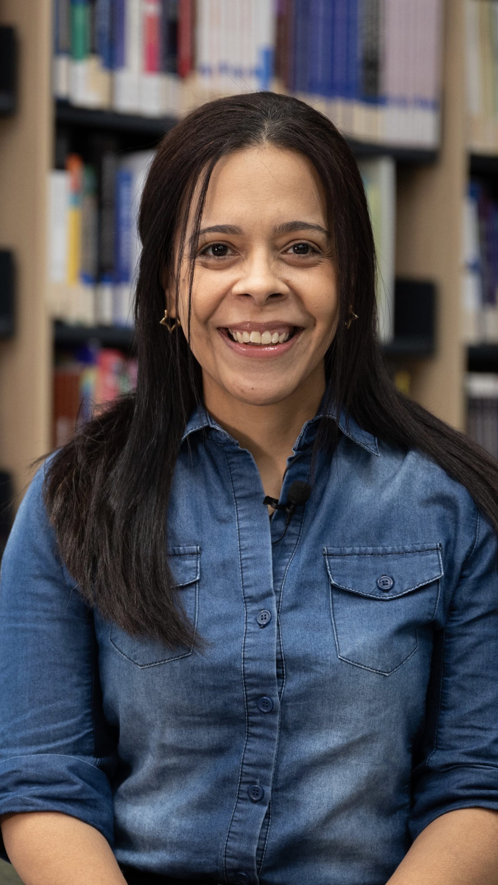 Person smiling and seated in a library setting, wearing a denim shirt with bookshelves in the background.