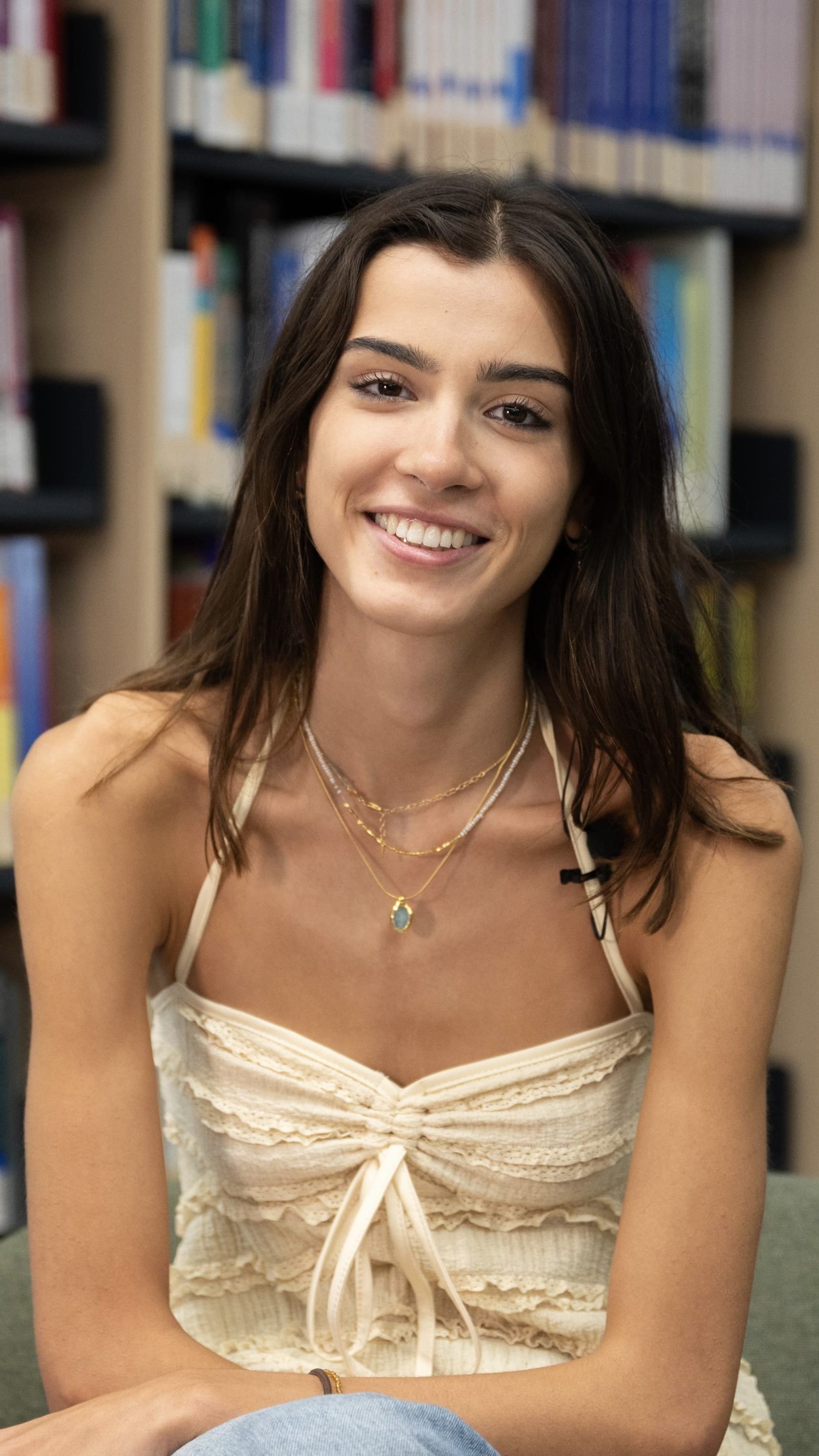 Person smiling while seated in a library, wearing a light cream top with layered detailing and gold necklaces. Bookshelves filled with colourful books are visible in the background.