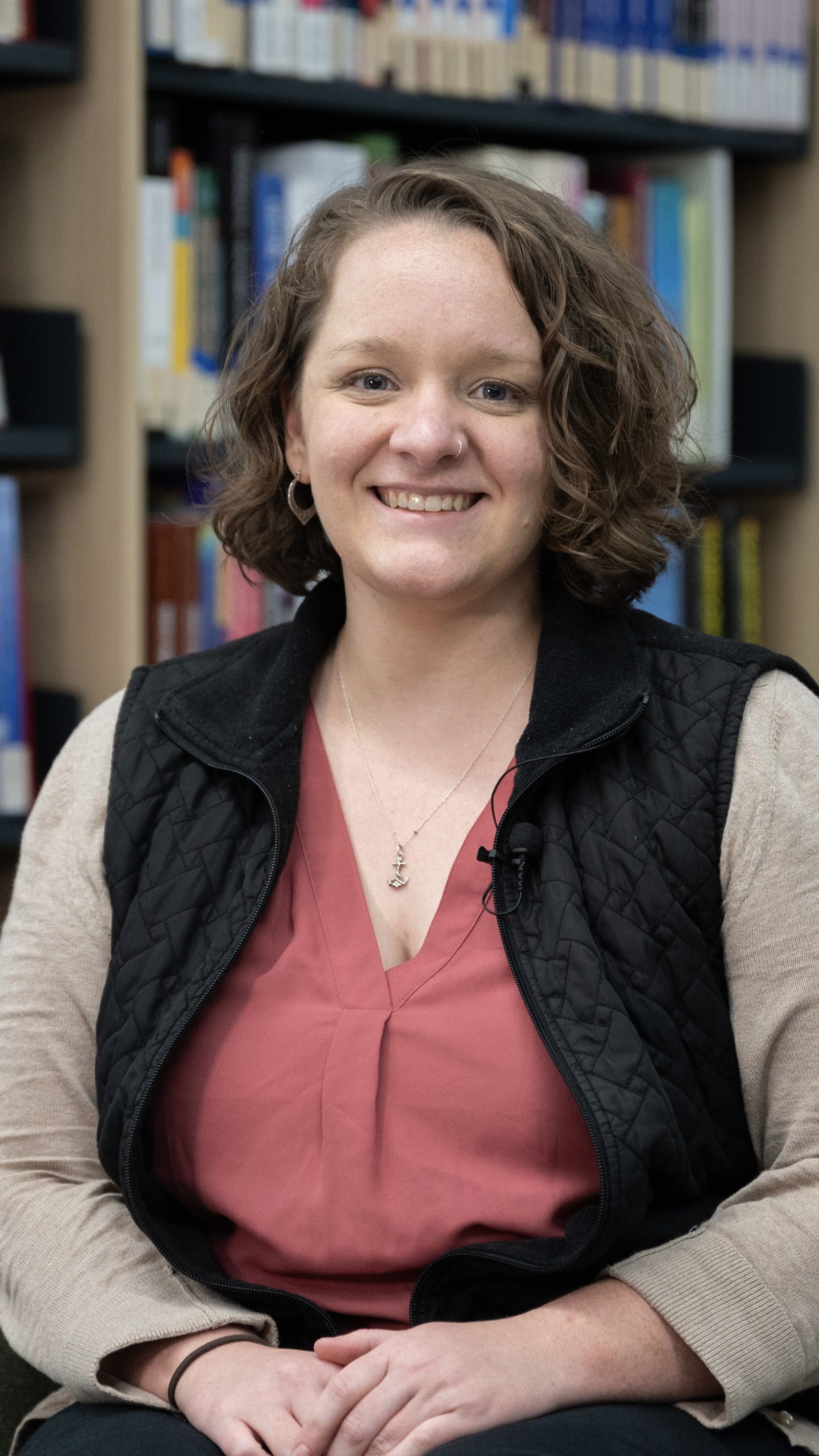 Person smiling while seated in a library, wearing a coral blouse with a black quilted vest and light cardigan. Bookshelves filled with colourful books form the background.