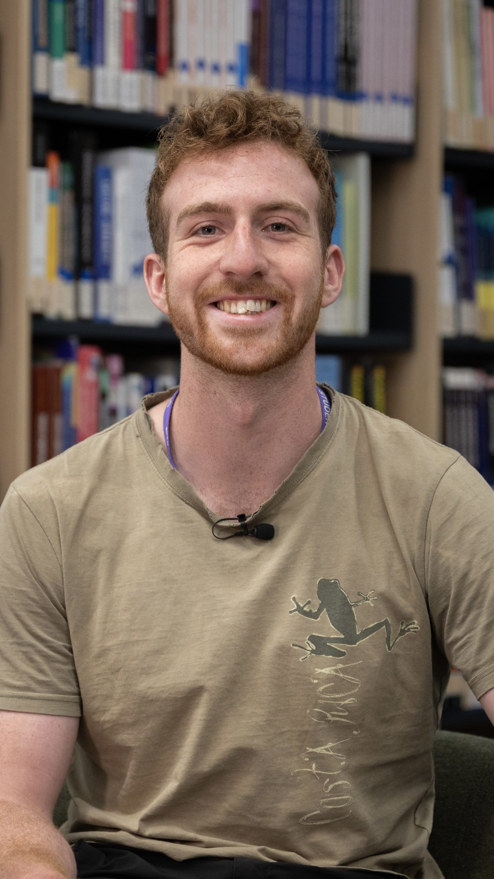 Person smiling while seated in a library, wearing a khaki T-shirt with a frog and “Costa Rica” design. Bookshelves filled with colourful books are visible in the background.