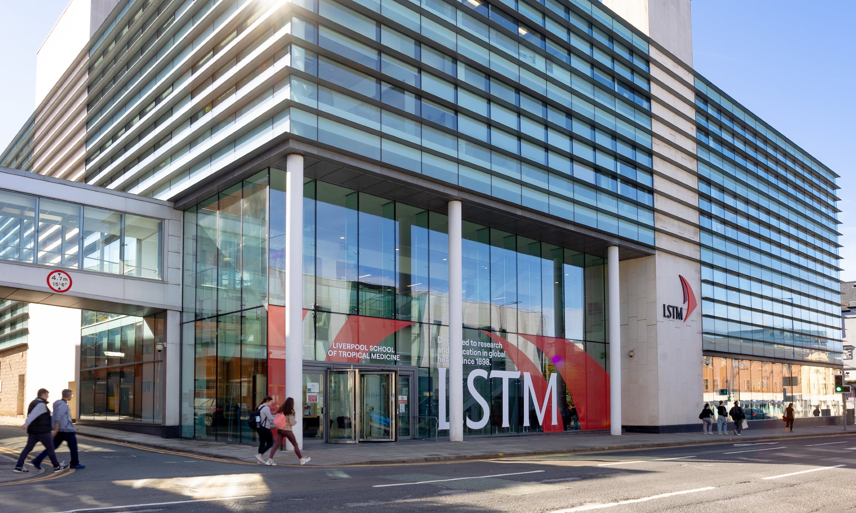 Exterior view of the Liverpool School of Tropical Medicine building on Pembroke Place, Liverpool, showing the glass-fronted entrance with the LSTM logo and people walking past on a sunny day.