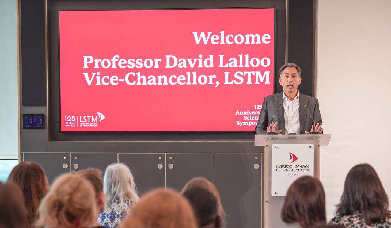 Professor David Lalloo, Vice-Chancellor of Liverpool School of Tropical Medicine, speaking at a podium during the 125th Anniversary Scientific Symposium. Behind him, a large red screen displays the welcome message and LSTM logo, while an audience listens attentively in the foreground.