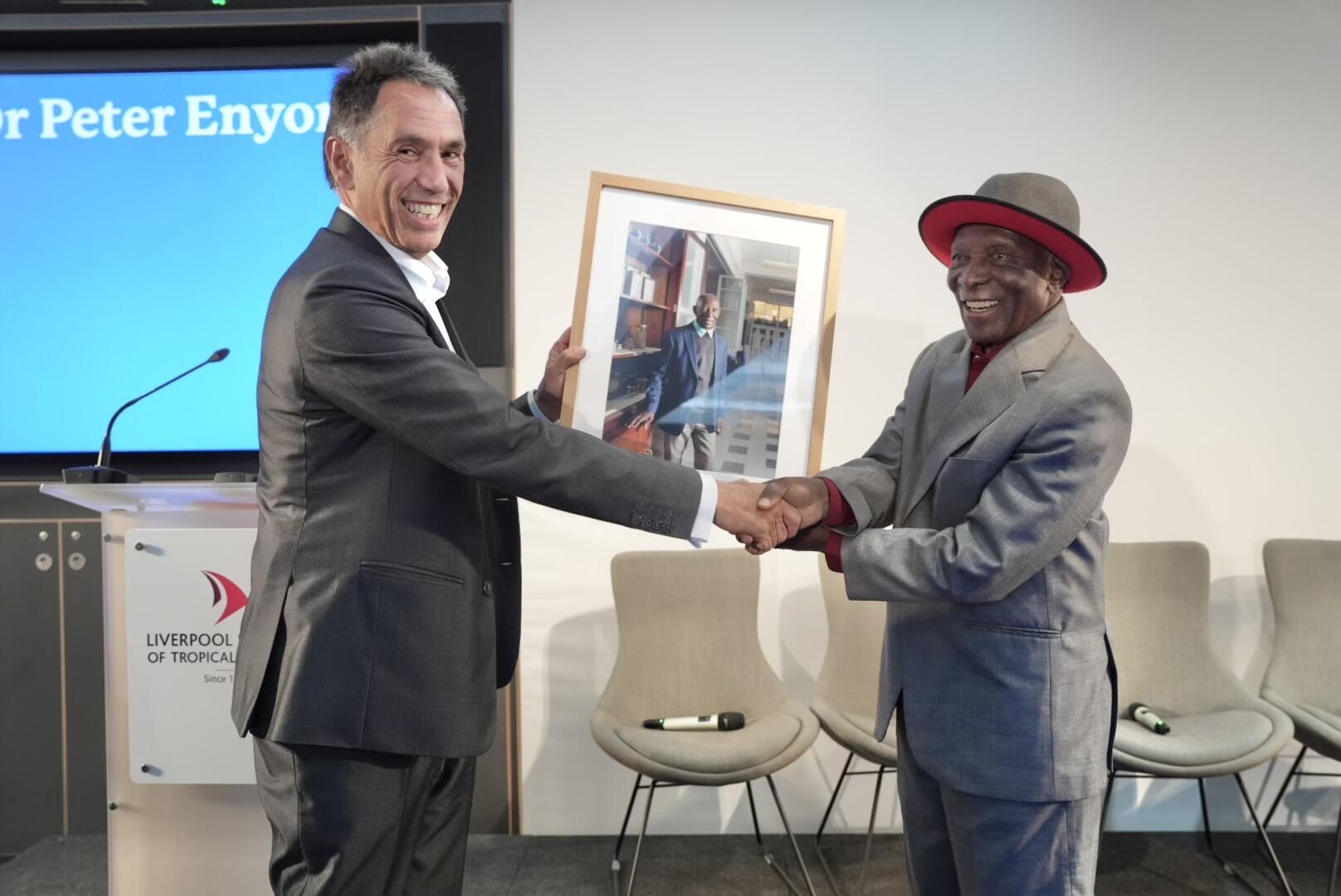 Two men in suits shake hands and smile on stage at the Liverpool School of Tropical Medicine. One holds a framed photograph of the other, presented as part of an award or recognition ceremony, with a presentation screen and chairs visible in the background.