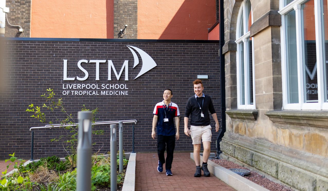 Two students walk and talk outside the Liverpool School of Tropical Medicine building, passing a wall displaying the LSTM logo and name on a sunny day.