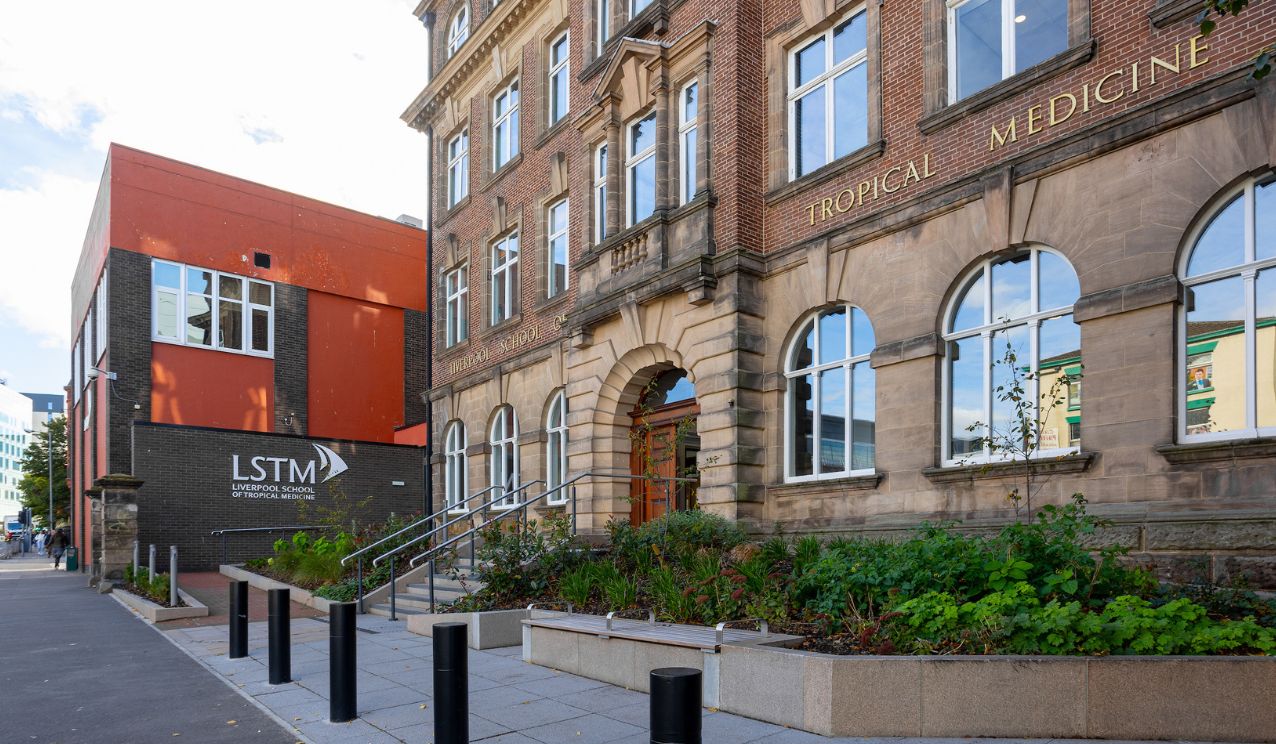 Exterior view of the Liverpool School of Tropical Medicine on Pembroke Place, showing the historic red-brick building alongside the modern LSTM extension with the school’s logo visible on the wall under a bright blue sky.