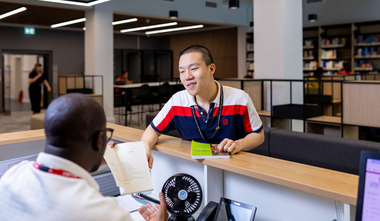 A student stands at a library help desk, smiling as they speak with a staff member. The staff member is holding an open book while the student holds two library books. Shelves of books and study areas are visible in the background.