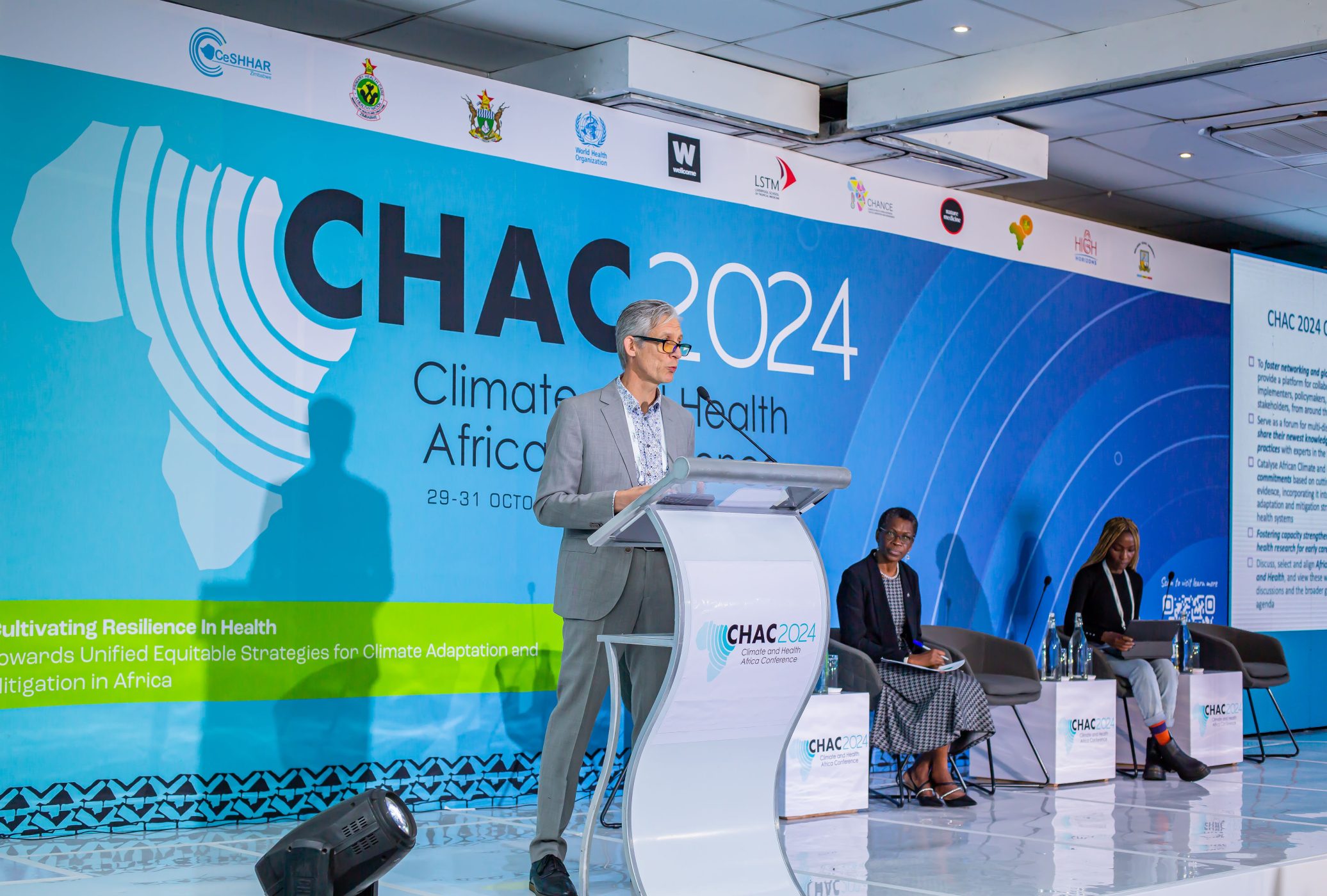 A speaker stands at a podium delivering a presentation at the Climate and Health Africa Conference 2024 (CHAC 2024), with two panelists seated on stage and a large blue backdrop displaying the event’s logo and theme.