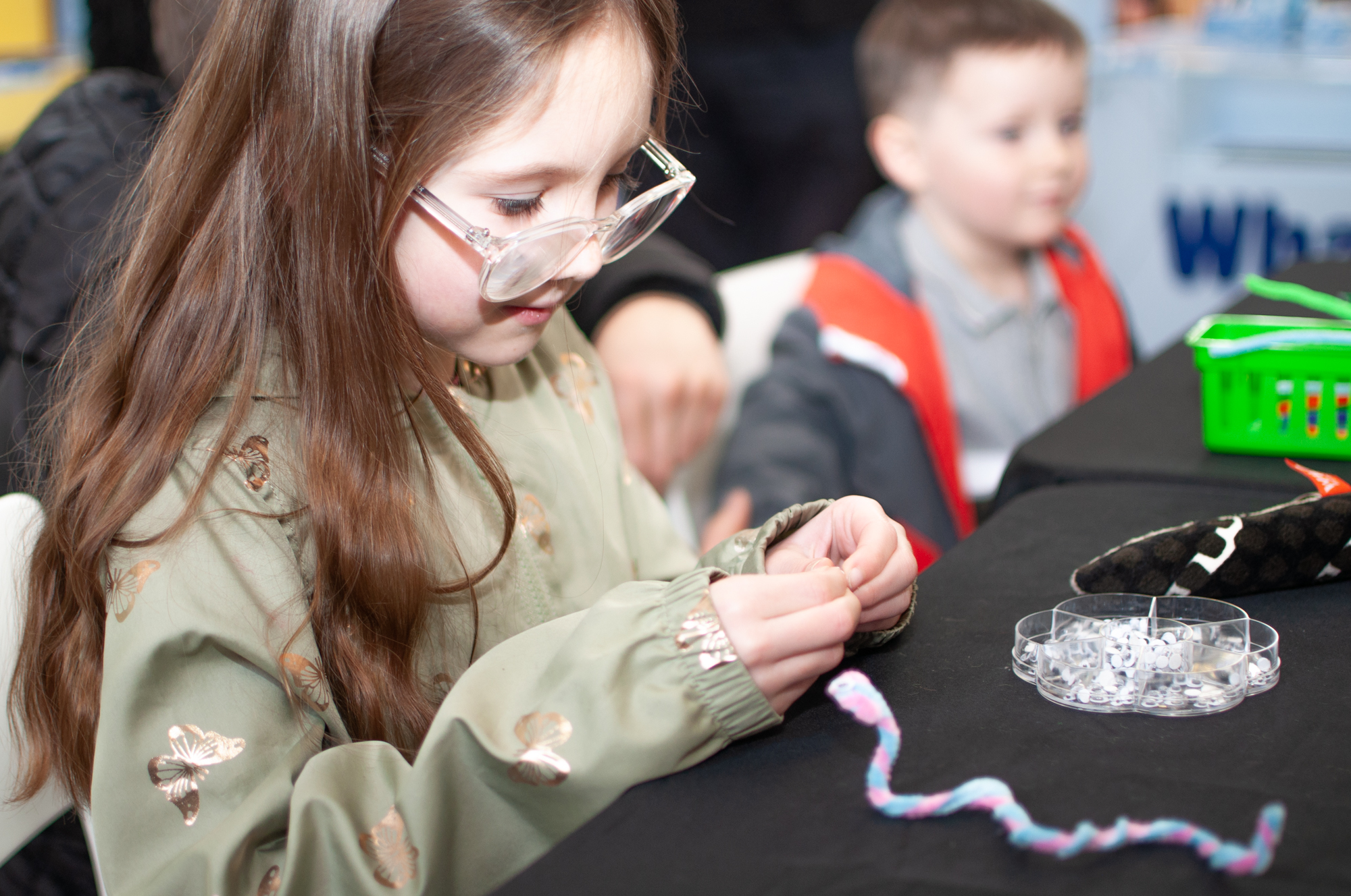 A young child with long brown hair and clear-framed glasses sits at a table, concentrating as they thread or inspect a small craft item in their hands. They wear a light green jacket with a butterfly print. In front of them are craft materials, including a tray of small googly eyes and a twisted, colourful pipe cleaner. Another child sits slightly out of focus in the background.