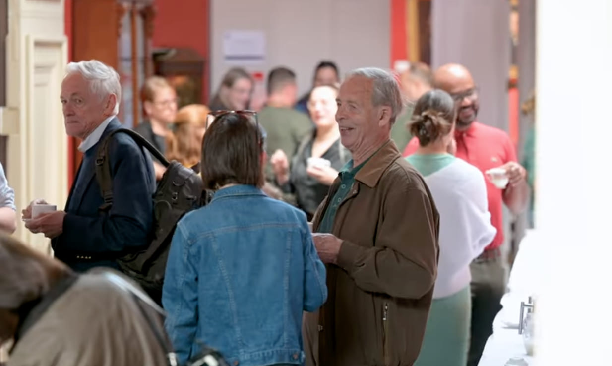 A group of people gather in a bright indoor space during an alumni event. Two older men chat and smile while holding drinks, surrounded by others engaged in conversation. The atmosphere is relaxed and sociable.
