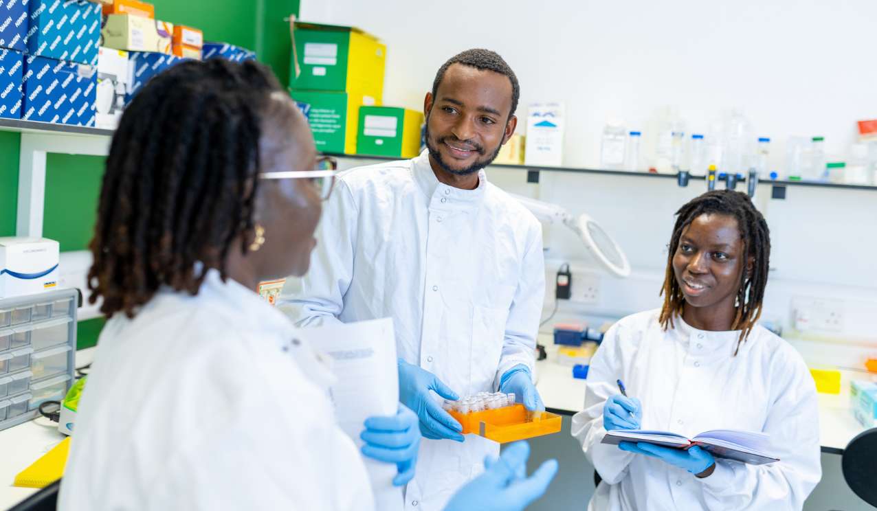 Three scientists in white lab coats talking in a bright laboratory, one holding a rack of sample tubes and another making notes in a notebook.
