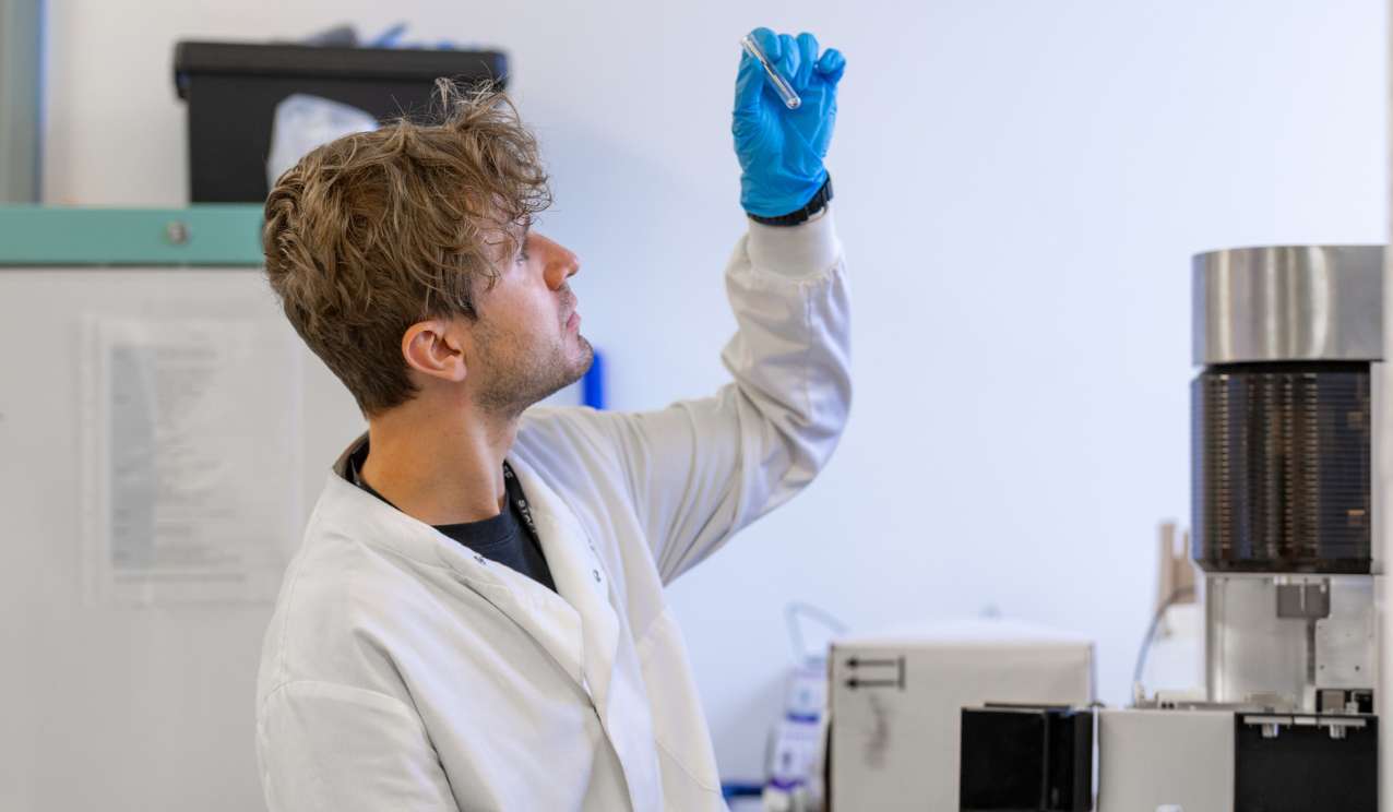 Scientist in a white lab coat and blue glove holding up a test tube to inspect it in a laboratory, with equipment visible in the background.