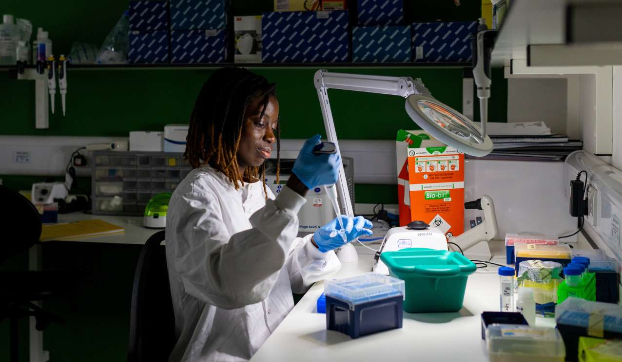 Scientist in a white lab coat and blue gloves using a pipette at a laboratory bench, surrounded by racks of tubes, equipment and a biohazard waste container under a task lamp.