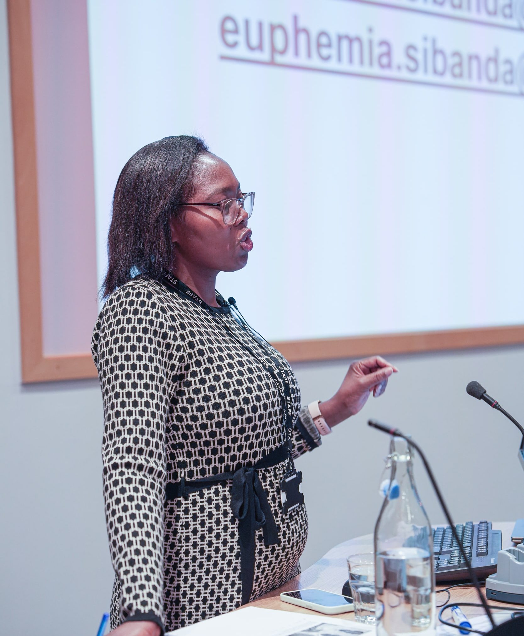 A woman in glasses speaks at a lectern in a lecture theatre, gesturing with one hand while a projected slide with text appears behind her and a microphone, papers and a water bottle sit on the desk in front.