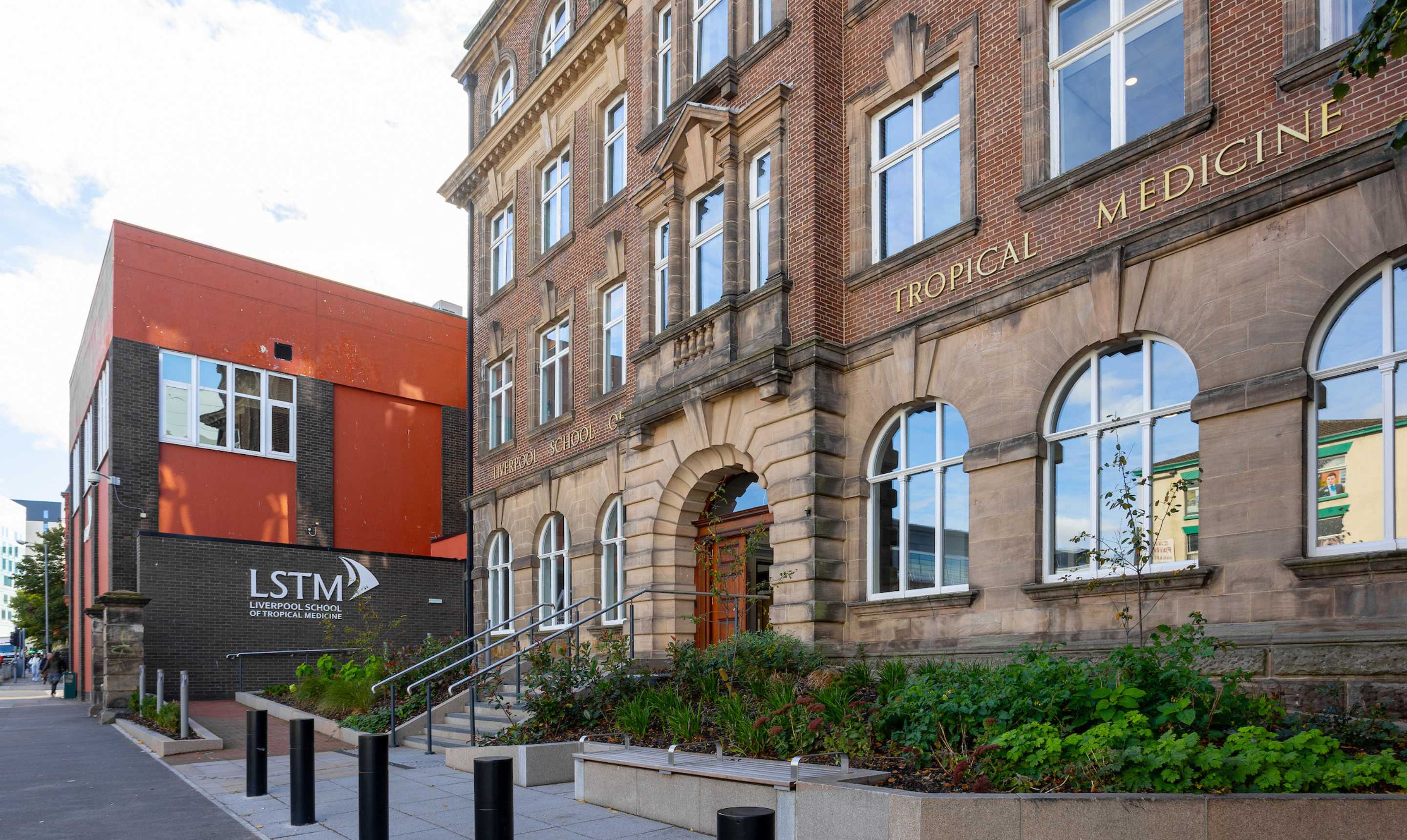 Liverpool School of Tropical Medicine’s Mary Kingsley building, with its red-brick façade and arched windows alongside a modern red annex displaying the LSTM logo, landscaped planting and an accessible ramp leading to the main entrance.
