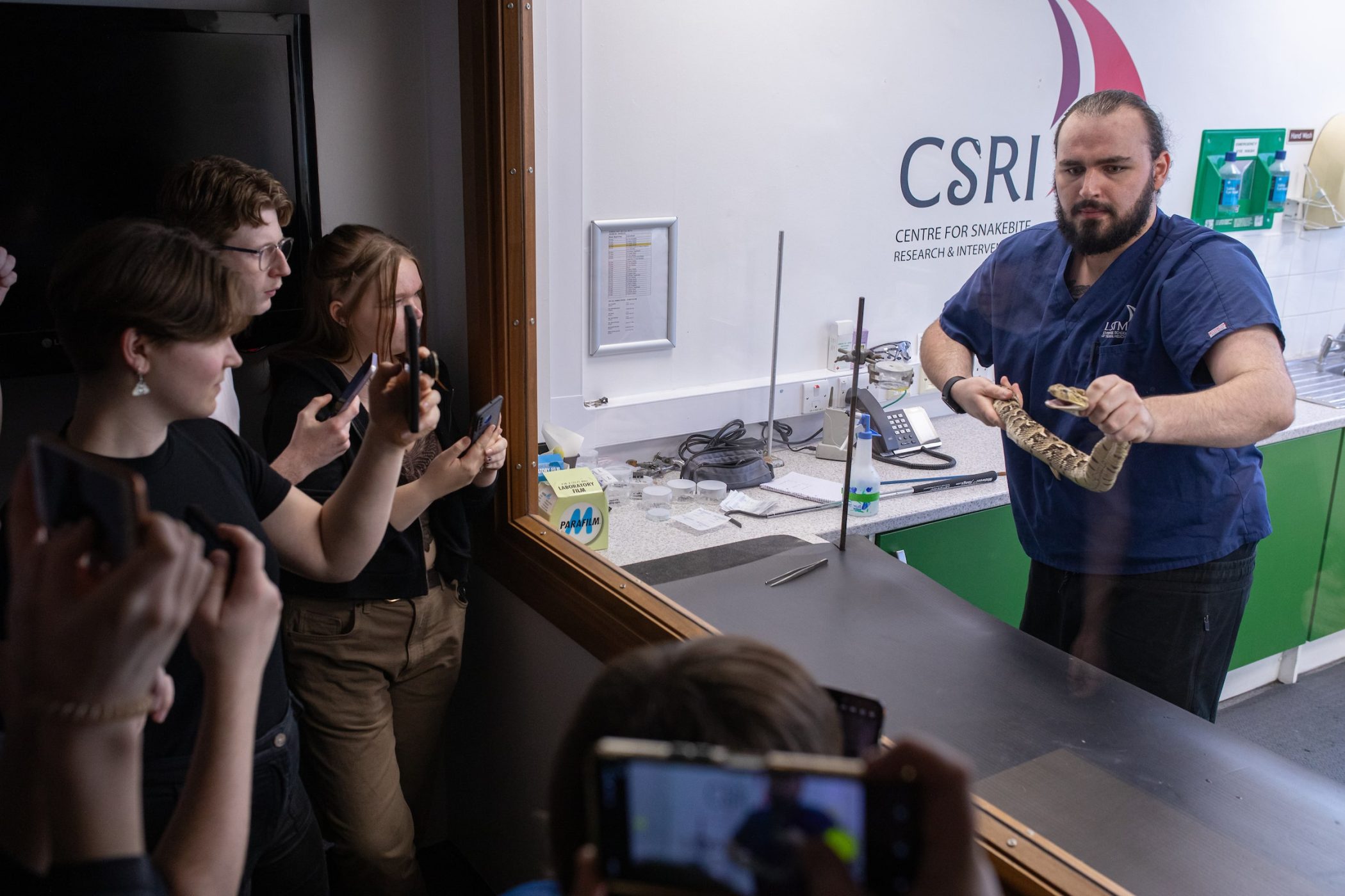 Students watch through a viewing window and film on their phones as a handler in blue scrubs holds a large snake in LSTM’s CSRI snakebite research laboratory, with the centre’s logo visible on the wall behind.